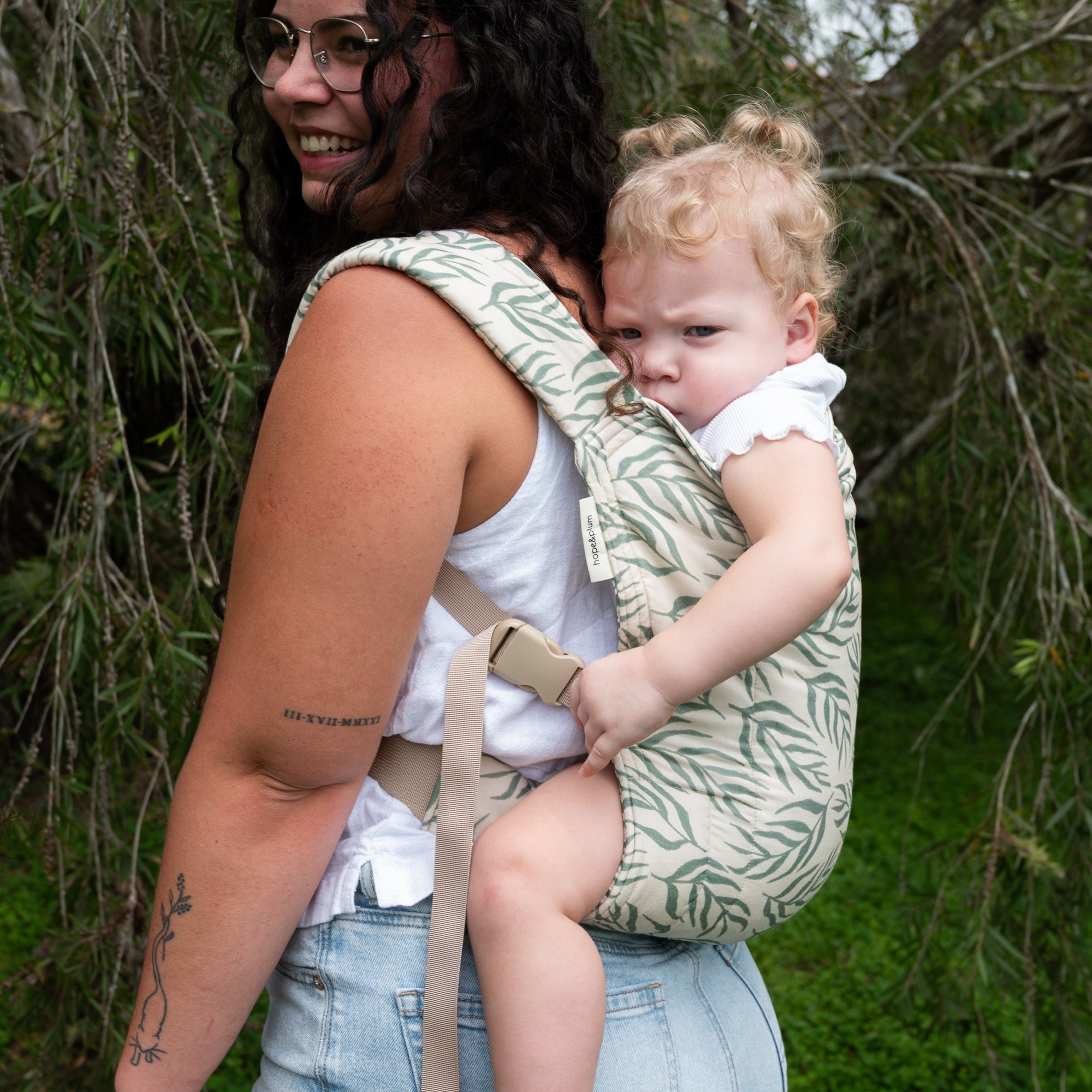 Woman carrying a baby in a leaf print baby carrier outdoors with greenery in the background