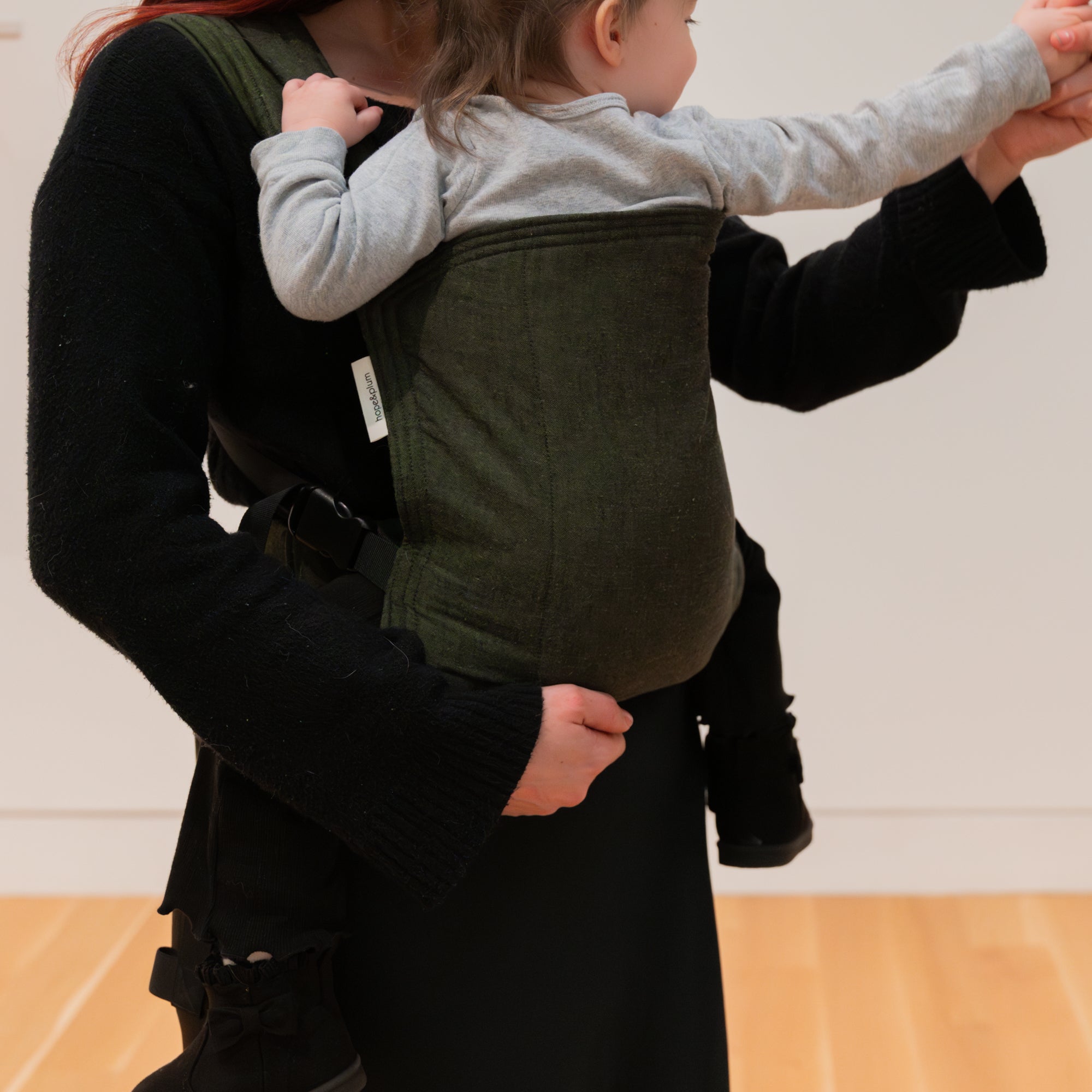 Person wearing a green baby carrier with a child inside, on a wooden floor.