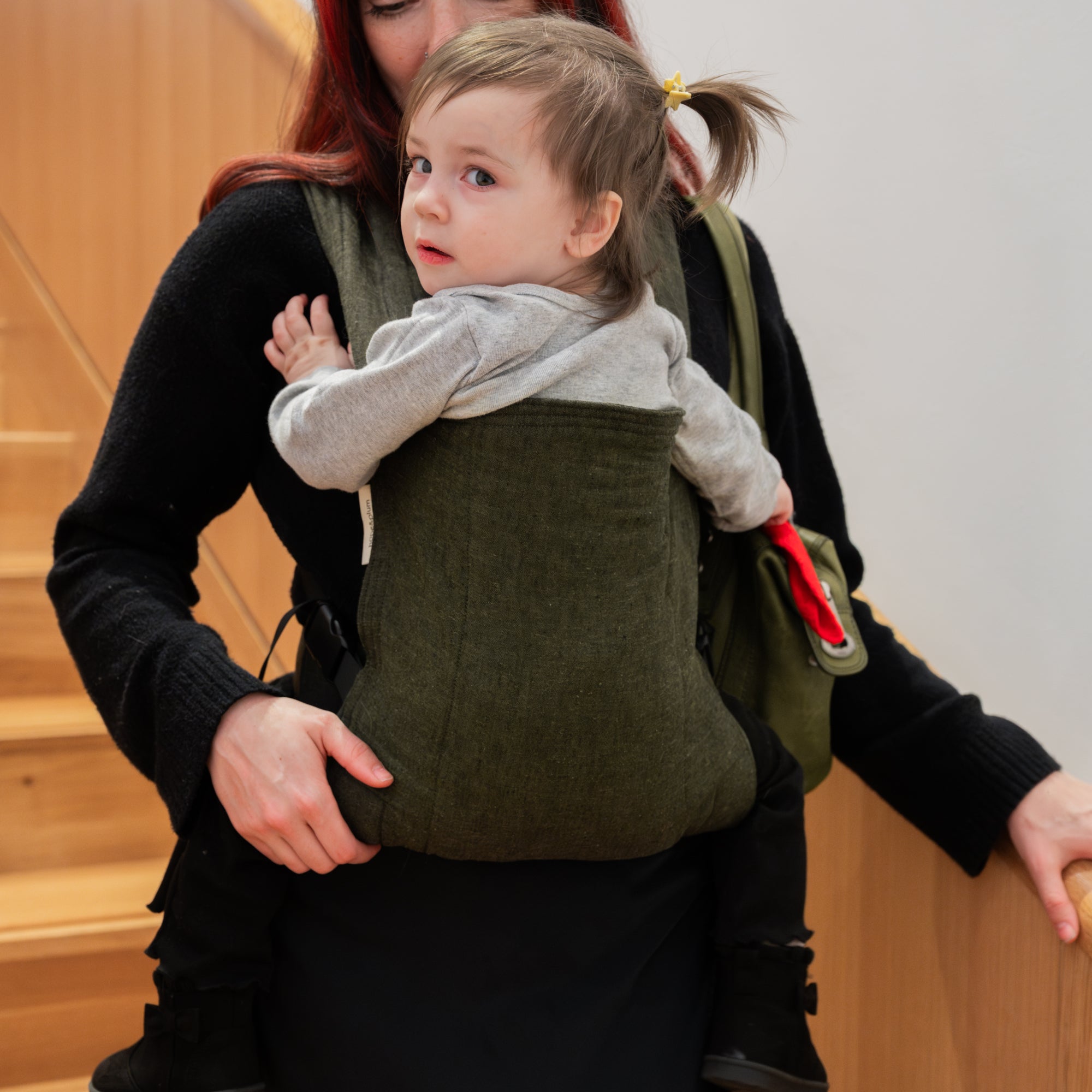 Woman holding a child in a green baby carrier indoors.