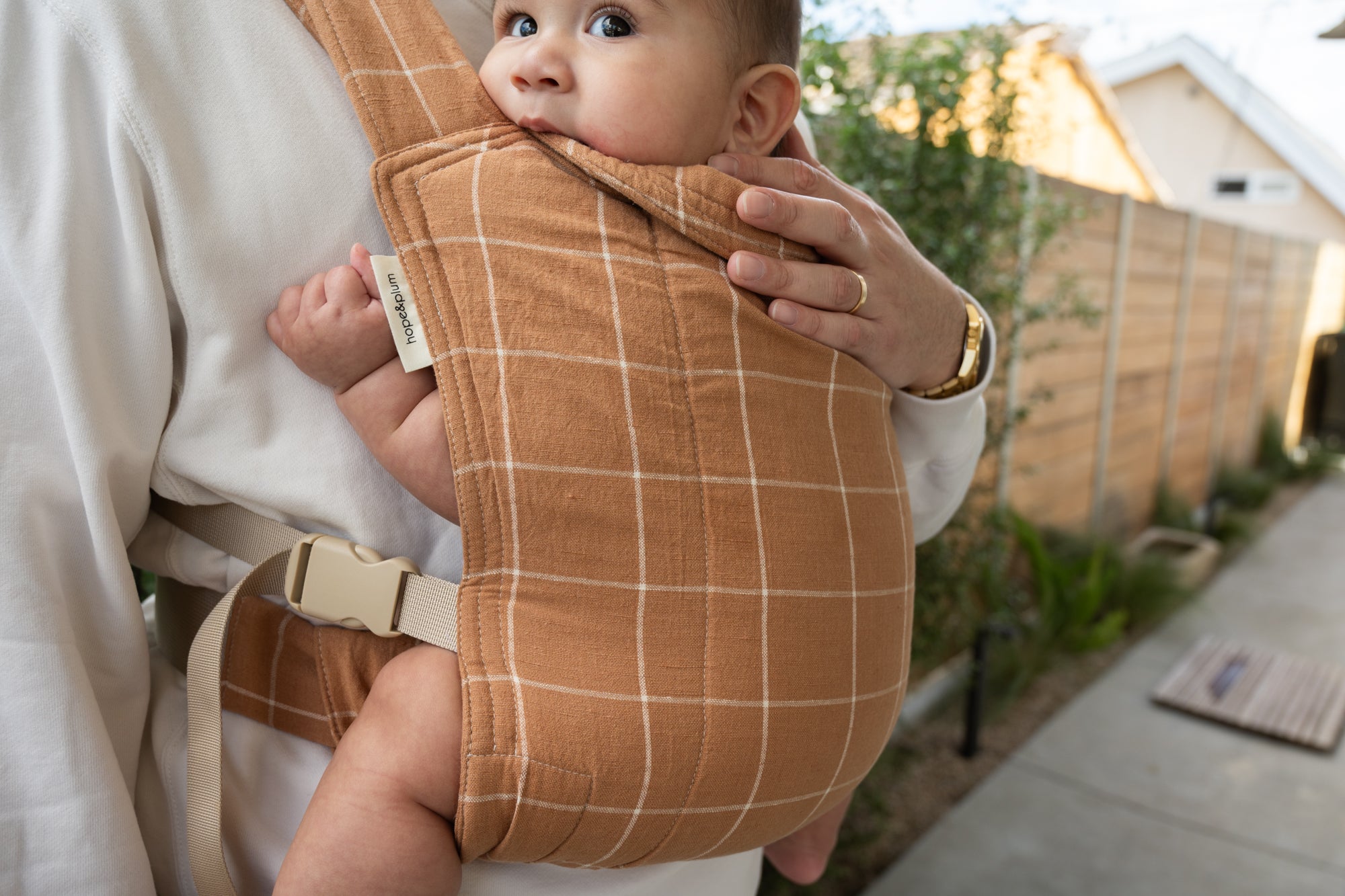 Baby in a brown checkered baby carrier held by an adult outdoors.