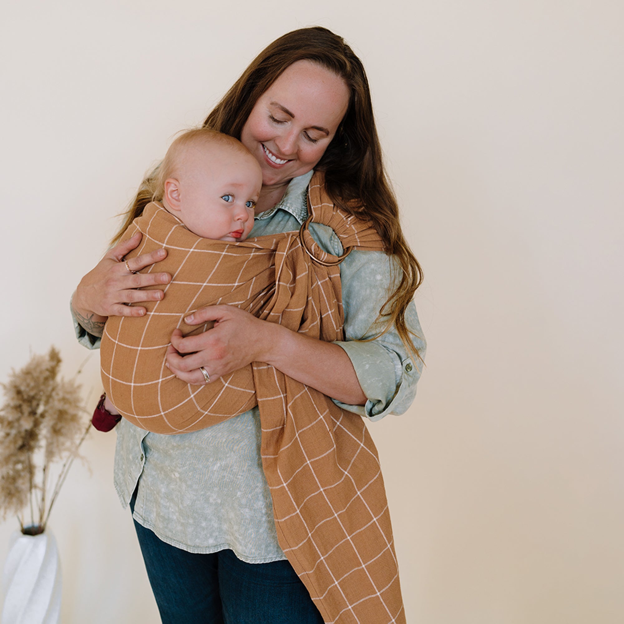 Woman holding a baby wrapped in a brown grid-patterned wrap against a plain background