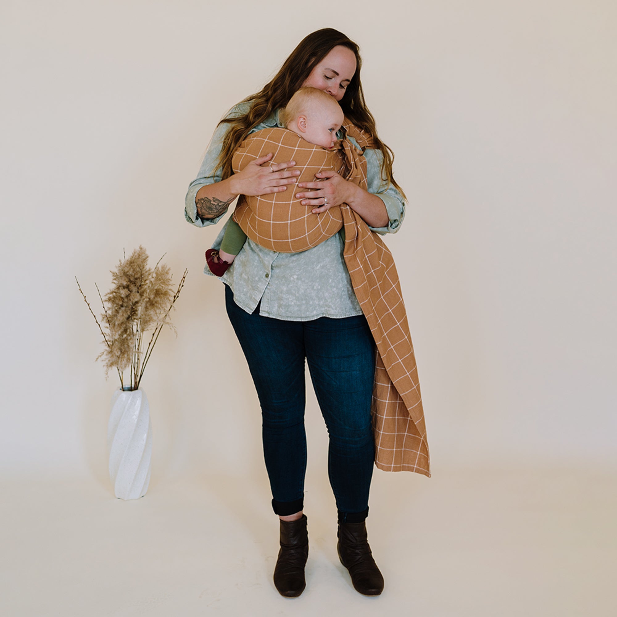 Woman holding a baby wrapped in a brown woven wrap against a plain background