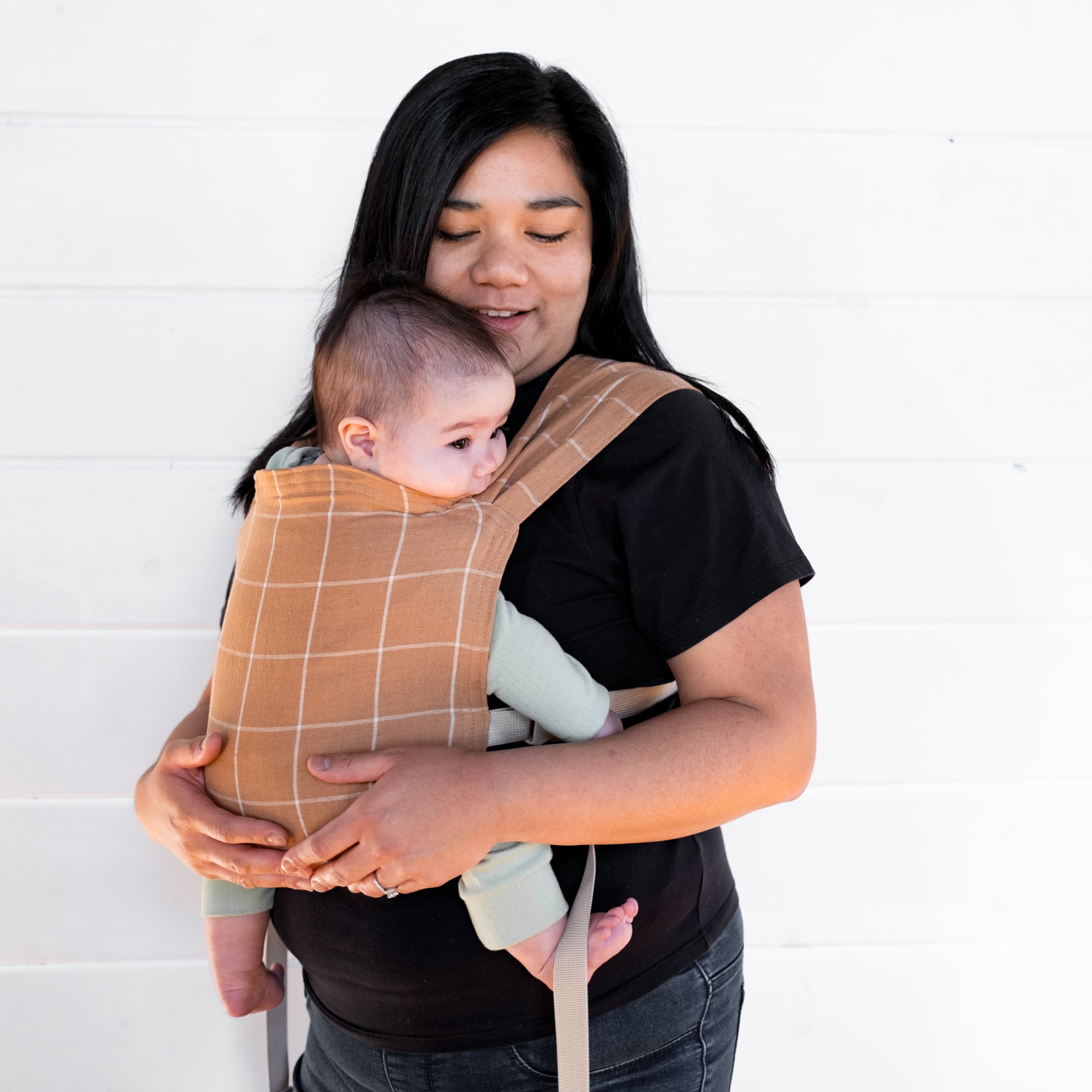 Woman holding a baby in a brown baby carrier with a white grid against a white background