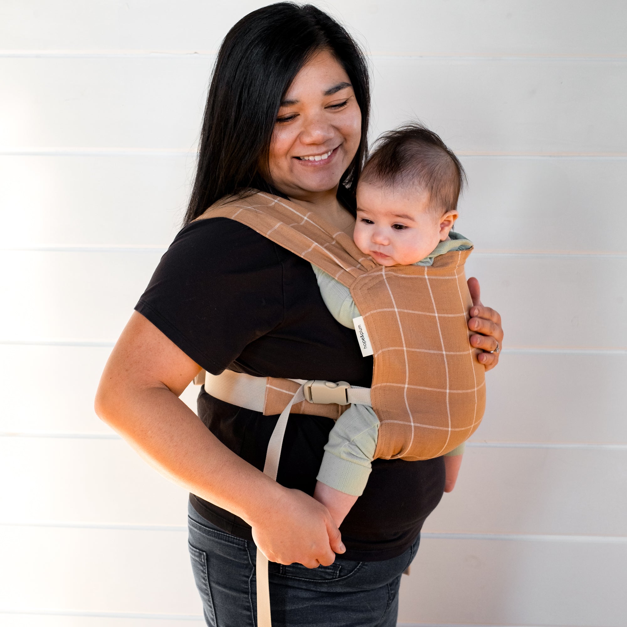 Woman holding a baby in a brown baby carrier with a white grid against a white background
