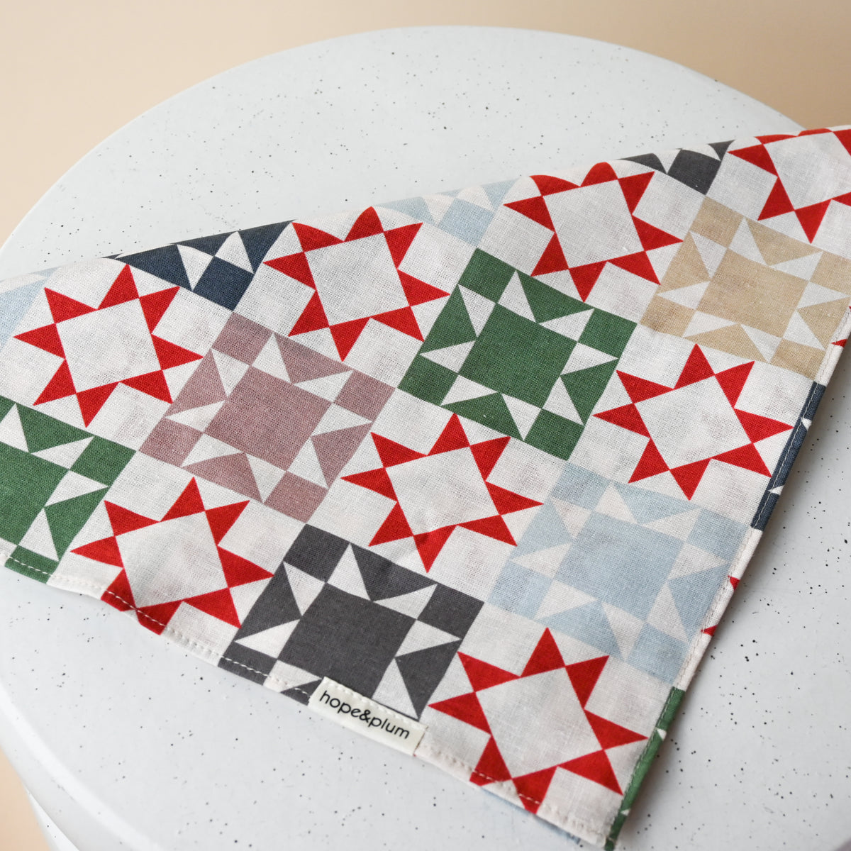 Large Bandana in Heirloom, folded on a white table, cream fabric printed with patchwork quilt star blocks in red, green, navy, slate blue and taupe.