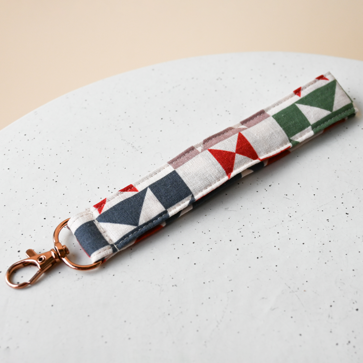 Key Fob in Heirloom, a fabric wristlet strap with a rose gold clasp, cream fabric printed with patchwork quilt star blocks in red, green, navy, slate blue and taupe.