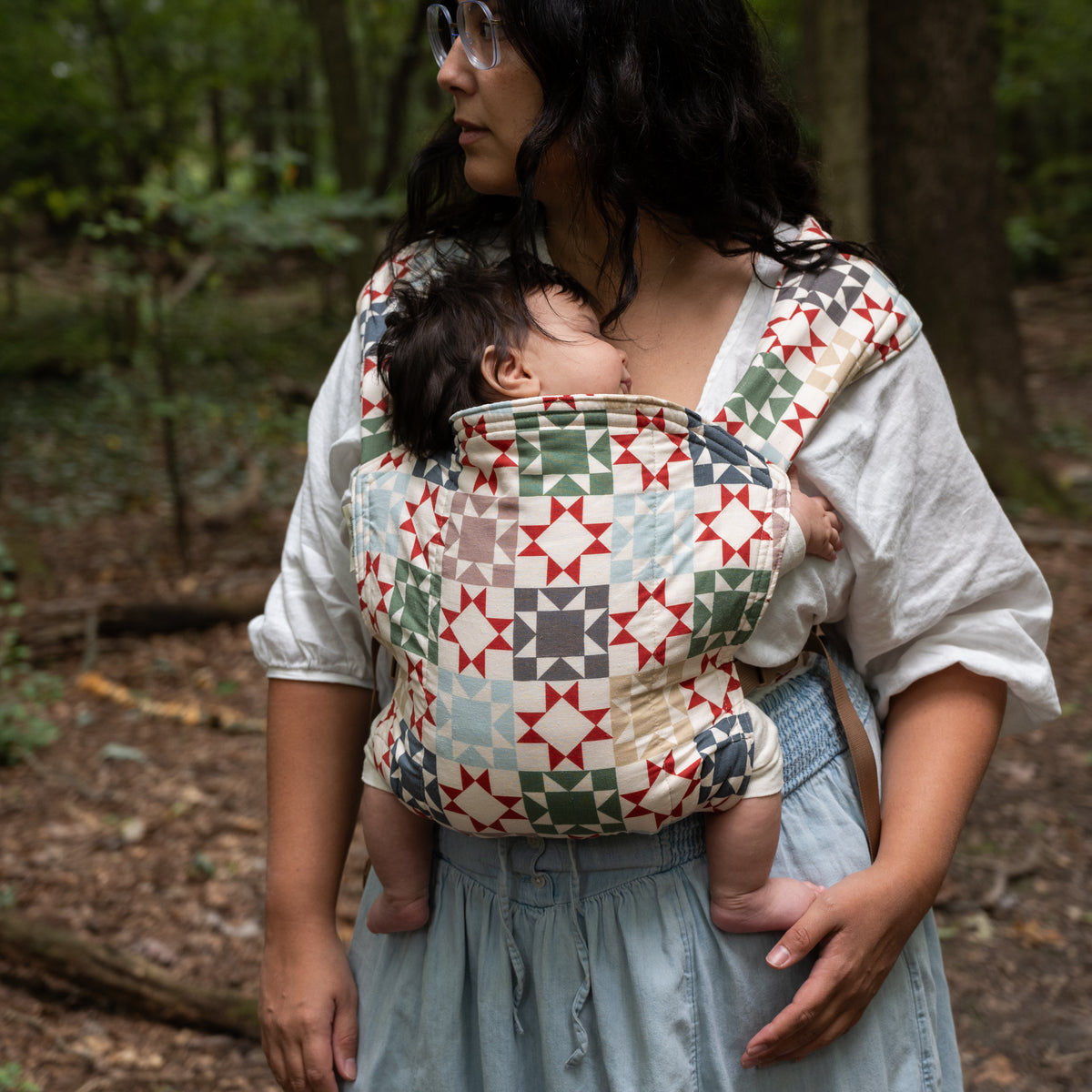 Heirloom Lark Baby Carrier worn in a front carry, a sleeping baby against the wearer's chest, in a patchwork quilt-block print of rust-red stars, sage and navy on warm cream.