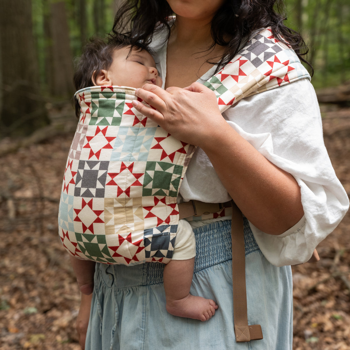 Close view of the Heirloom Lark Baby Carrier body panel and tan waist strap, a hand cupping the sleeping baby's head, in rust-red, sage and navy quilt blocks on cream.
