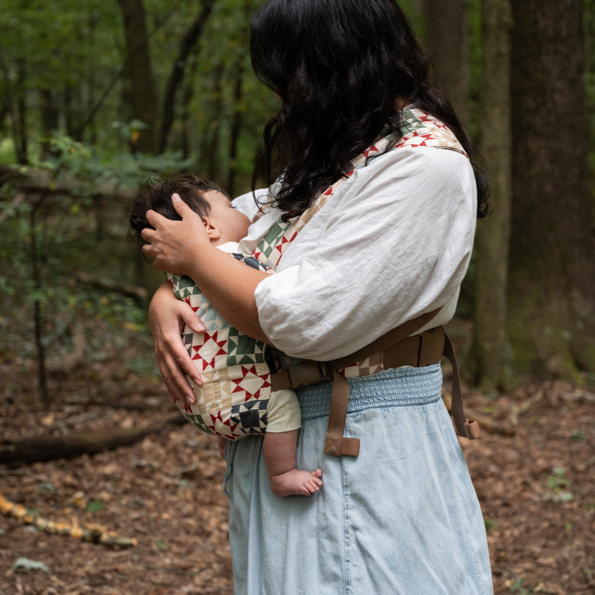Side view of the Heirloom Lark Baby Carrier in a front carry, the padded waistband and tan straps visible, the baby's legs supported in a seated position.