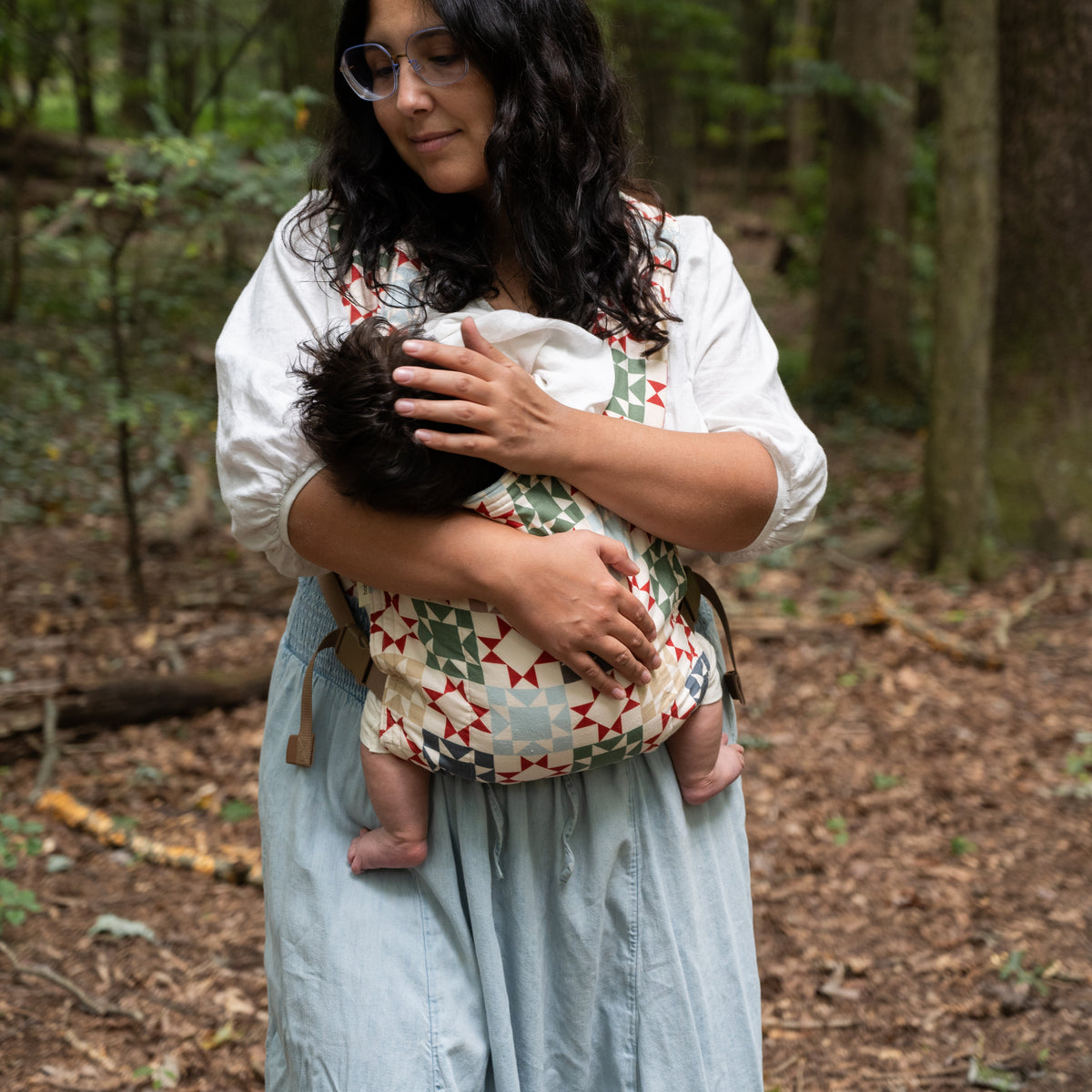 A wearer looks down at the baby carried in the Heirloom Lark Baby Carrier, the quilt-block panel of rust-red stars and sage on cream across the front.