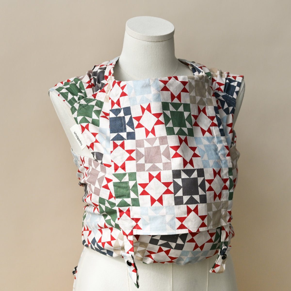Heirloom Meh Dai carrier shown on a dress form, cream fabric printed with patchwork quilt star blocks in red, green, navy, slate blue and taupe, with the hope&plum label on the strap.