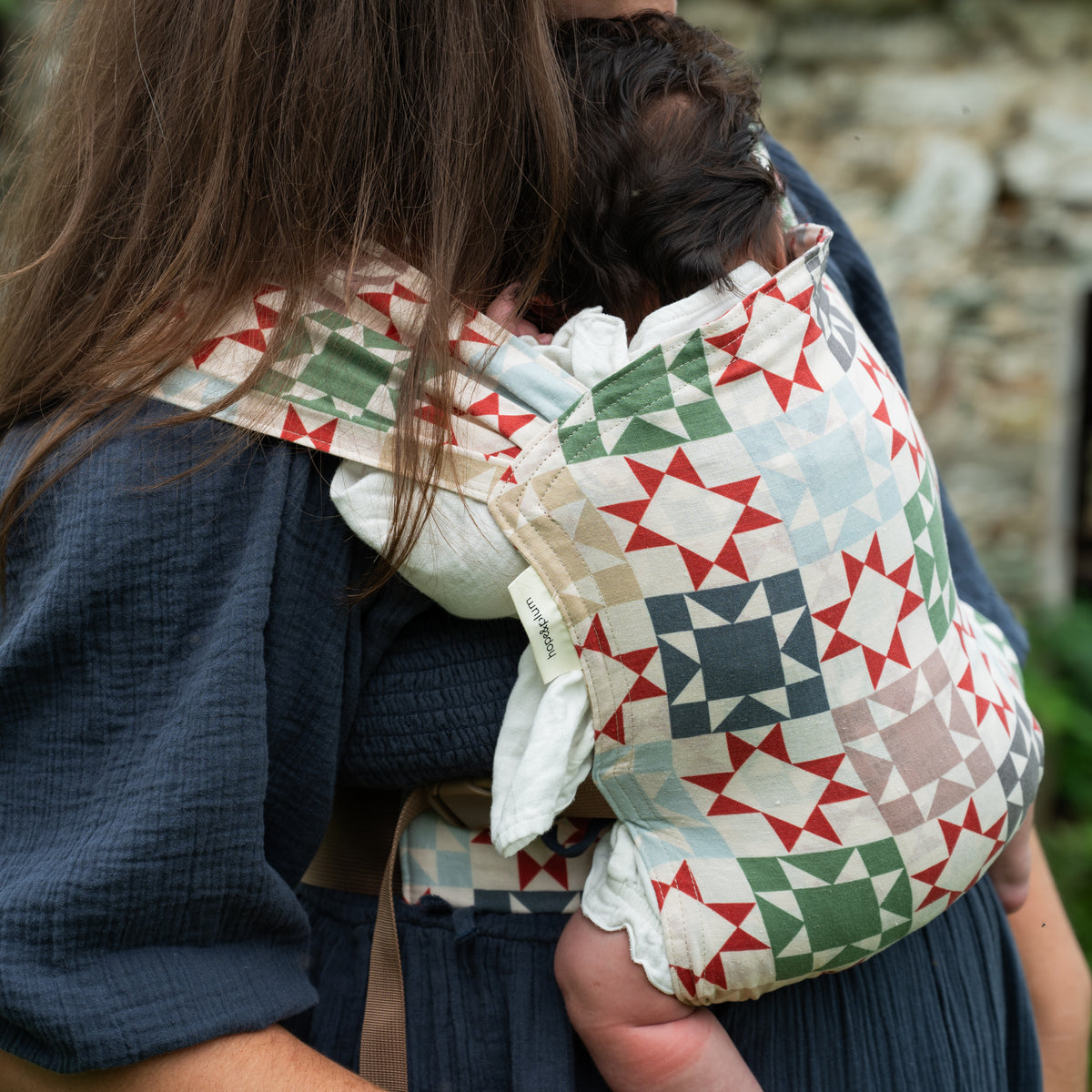 A newborn rests against the wearer's chest in the Heirloom Sprout Newborn Carrier, seen over the shoulder, showing star blocks in rust-red, sage, navy and seafoam on cream.