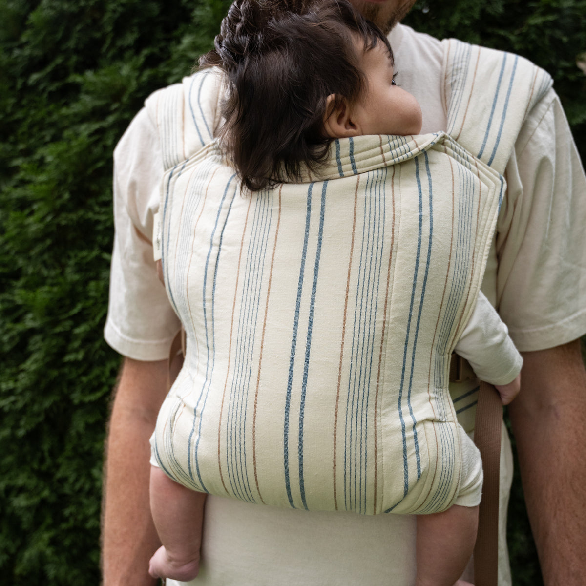 A baby rides in a back carry in the Lugano Lark Baby Carrier, the full striped panel of navy and tan on cream supporting them.