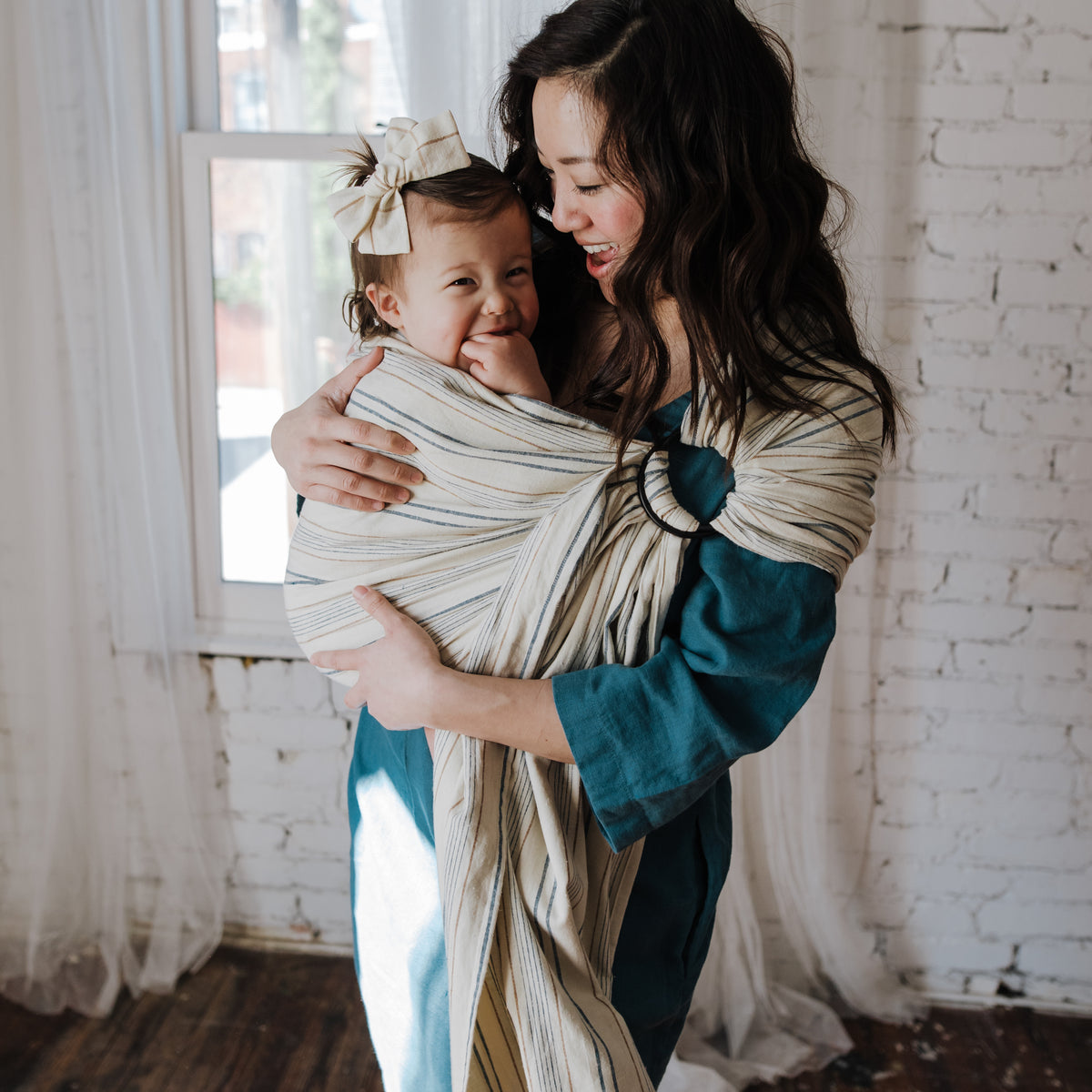 A baby is carried upright in the Lugano Ring Sling indoors, the striped chambray gathered through dark rings at the wearer's shoulder.
