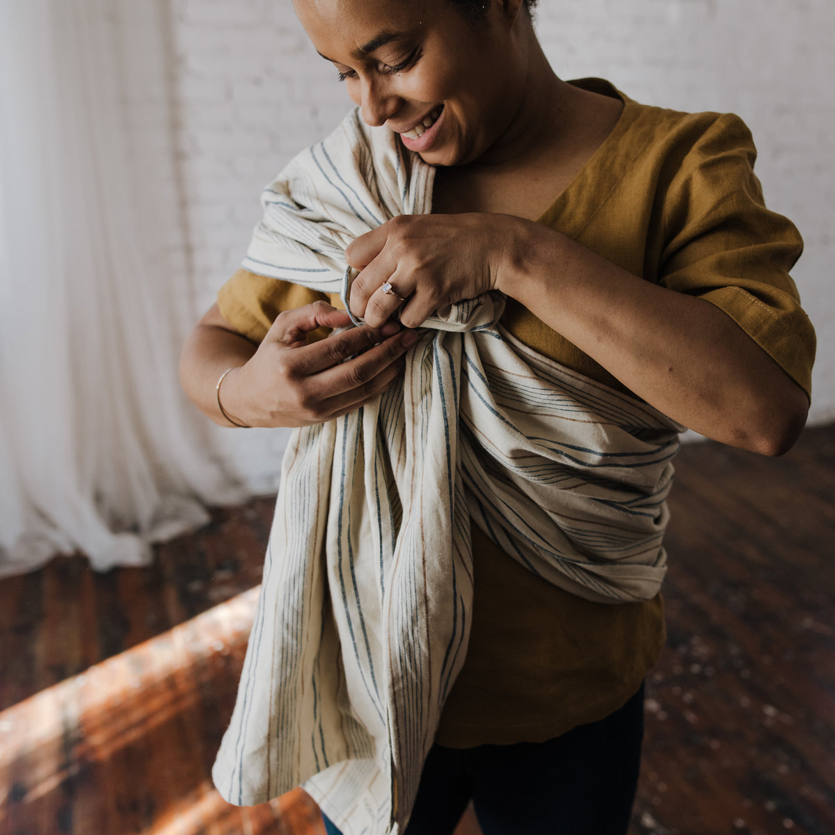 A wearer adjusts the tail of the Lugano Ring Sling across their shoulder, showing the drape of the woven chambray in navy and tan stripes on cream.