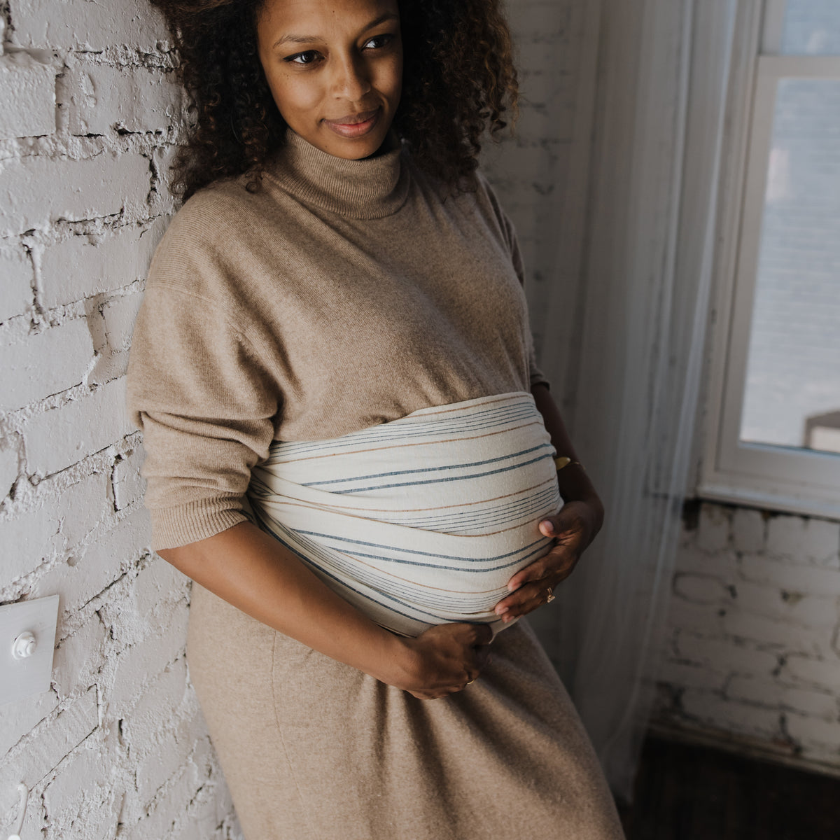 A wearer stands with the Lugano Ring Sling across the body, the woven navy and tan stripes on cream gathered into a smooth band.