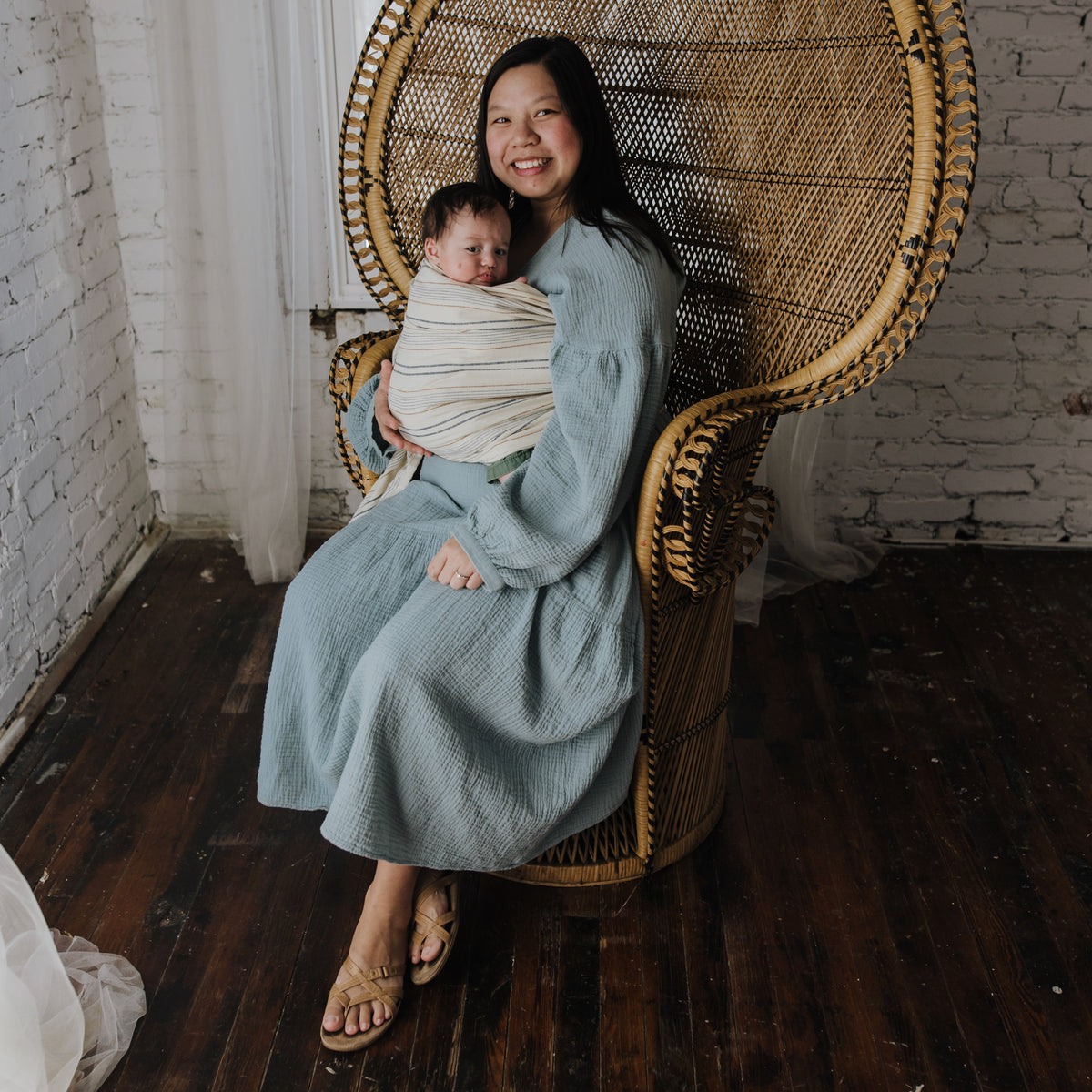 A wearer sits in a rattan chair with a newborn in the Lugano Ring Sling, the striped chambray drawn across the body.