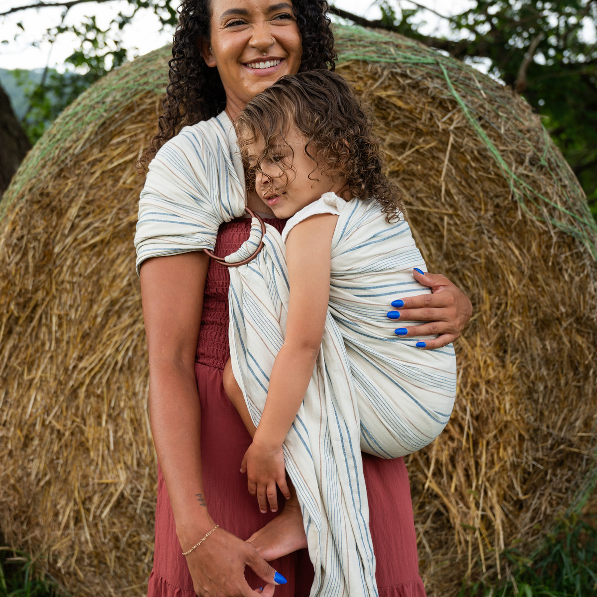 A smiling wearer carries a curly-haired toddler in the Lugano Ring Sling outdoors, the bronze rings at the shoulder above woven navy and tan stripes on cream.