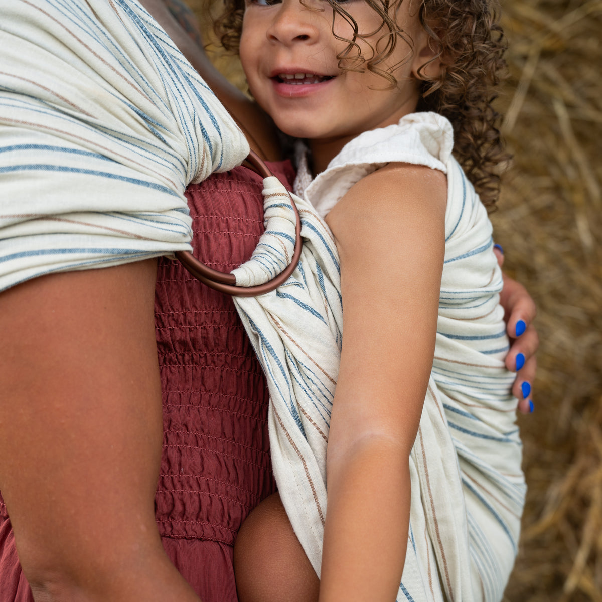 Close view of the Lugano Ring Sling's bronze rings with the striped tail threaded through, showing the woven navy and tan stripes on cream chambray.