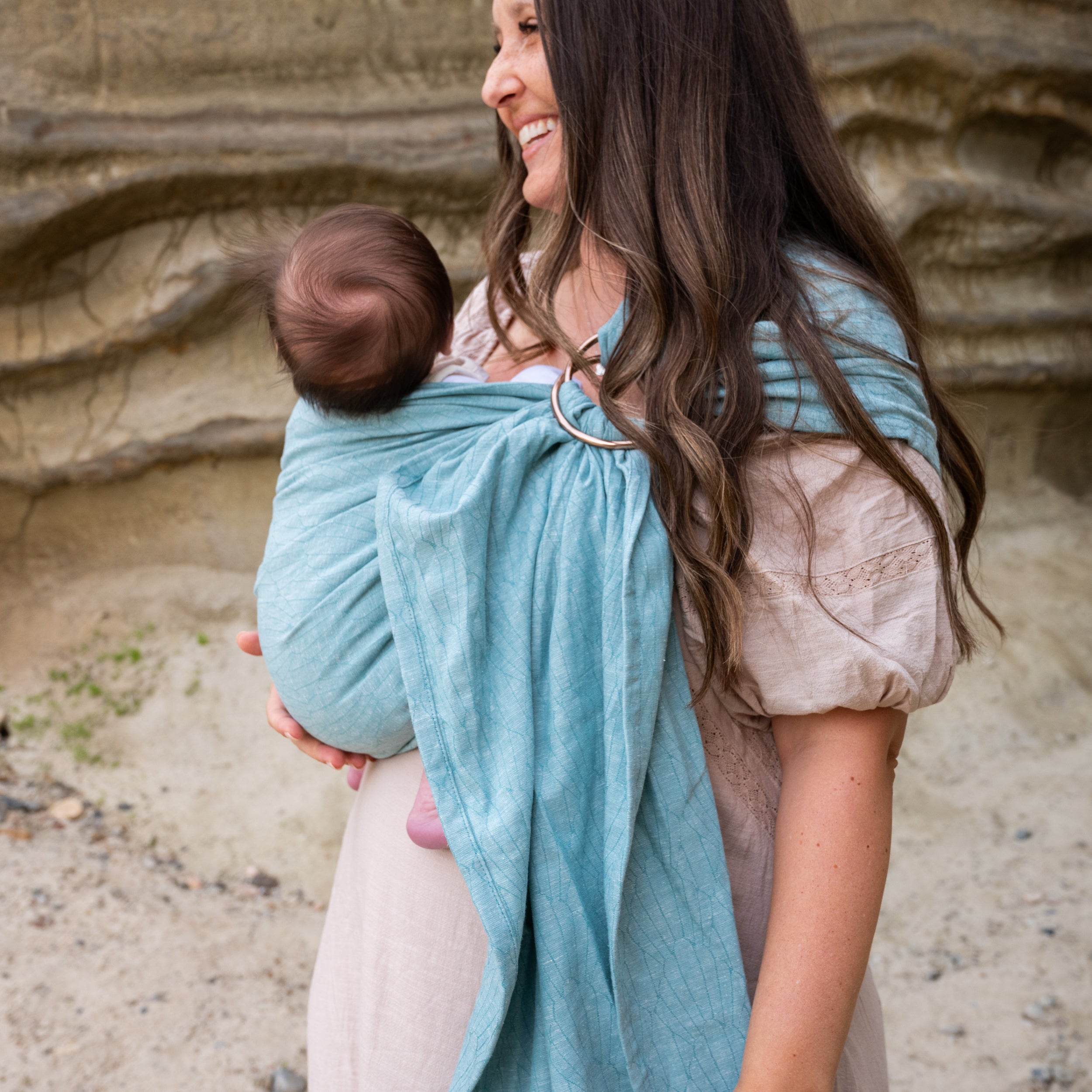 brunette woman wearing her infant in a blue seashell woven print ring sling against a sandy background