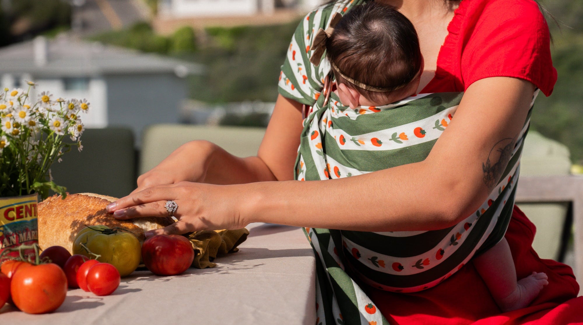 A close up of a woman in a red dress sitting at a table cutting bread, wearing her baby in a green and white ring sling with tomatoes and tulips on it.