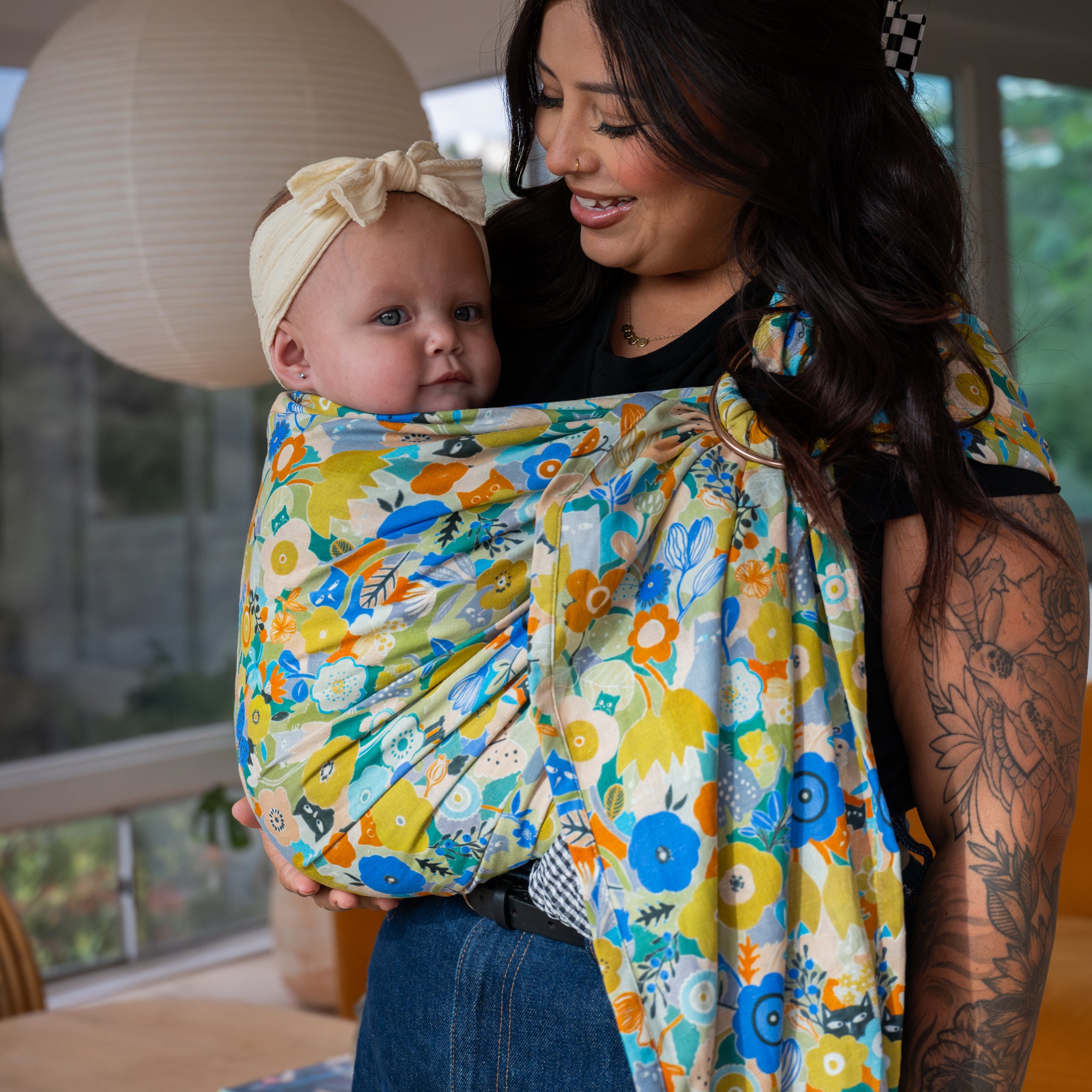 Woman holding a baby wrapped in a colorful floral ring sling in a home setting
