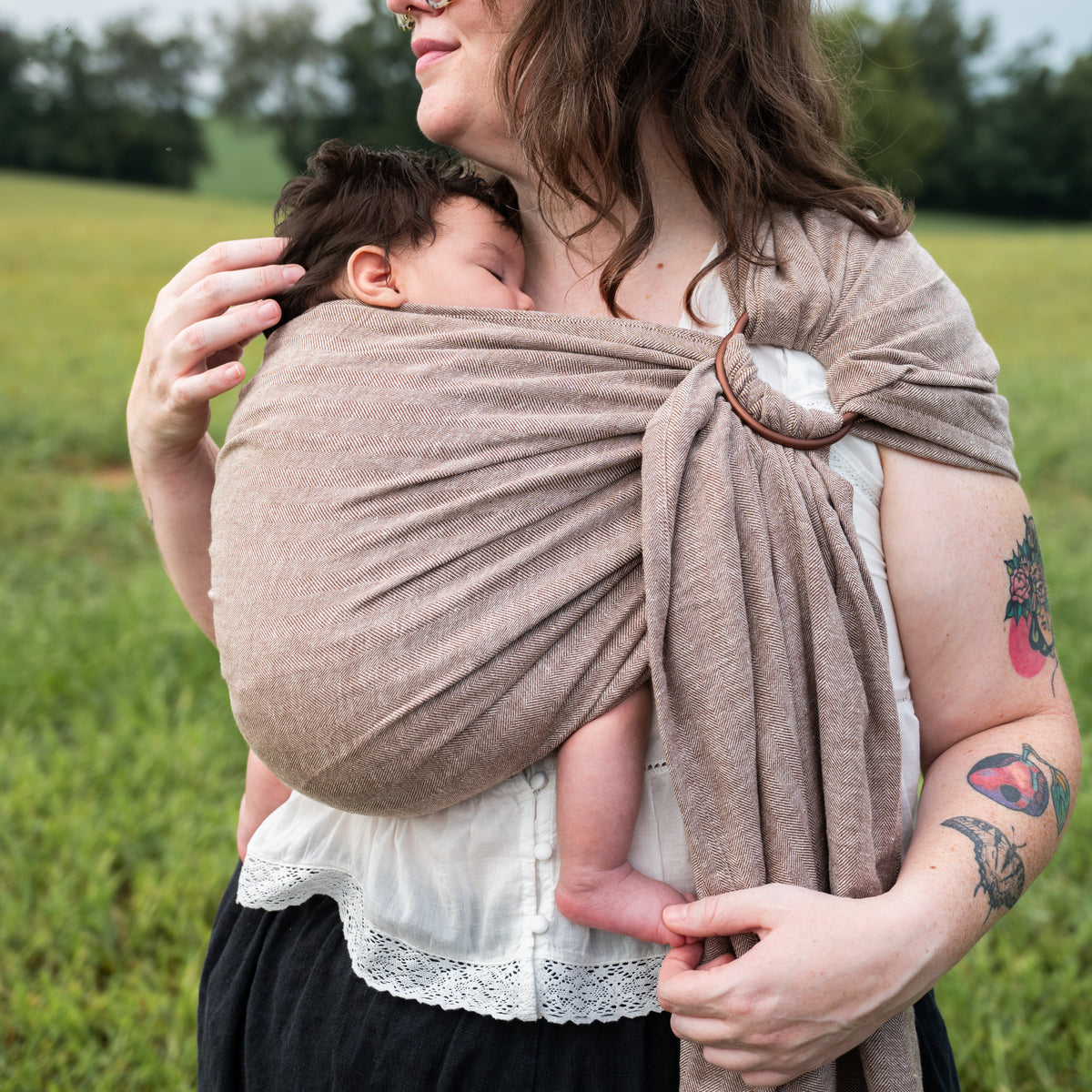 Close-up of a woman holding a sleeping newborn wrapped in the Oak Ring Sling's chocolate and oatmeal herringbone weave, showing the shoulder ring.