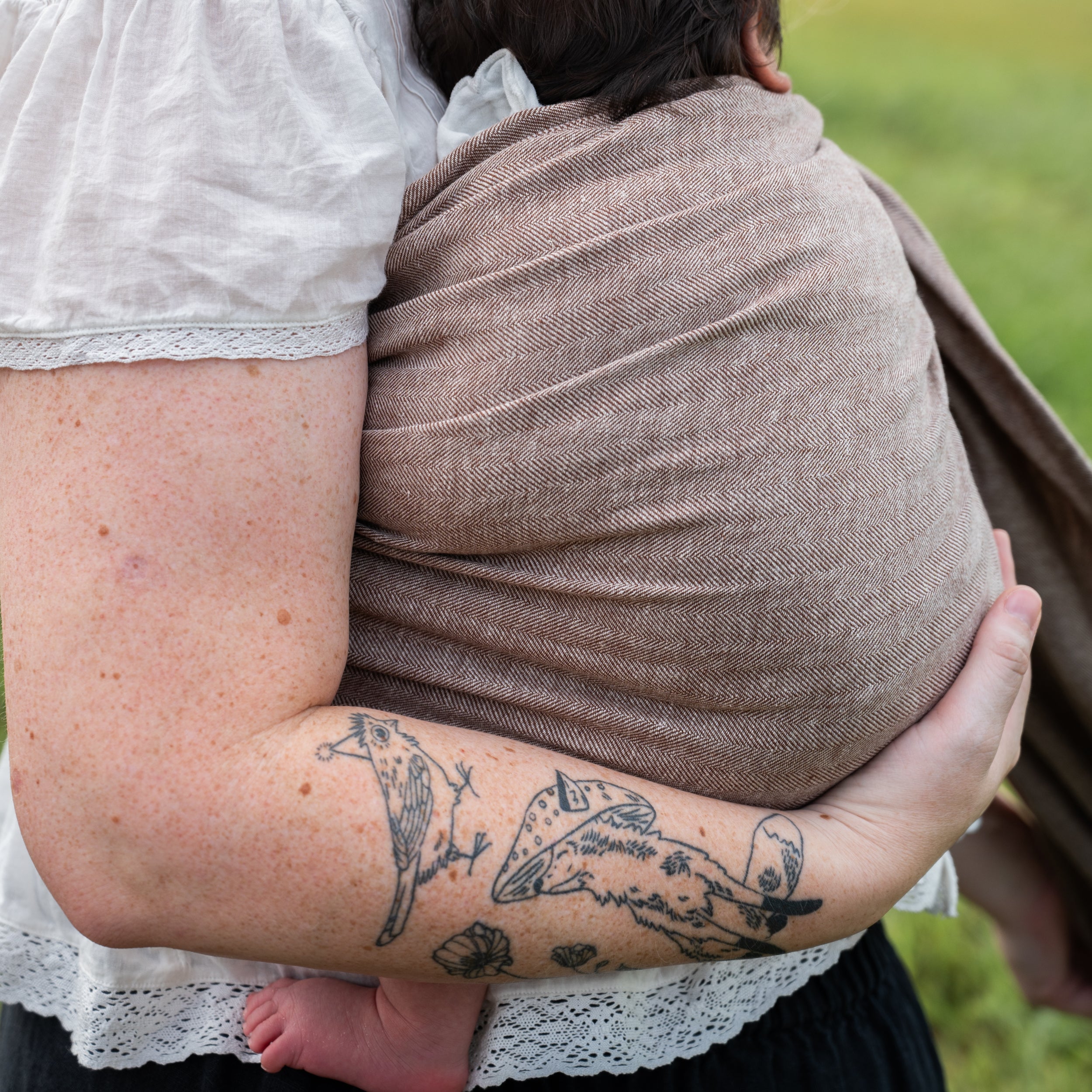 Close-up of the Oak Ring Sling's chocolate and oatmeal herringbone weave wrapped around a woman's back while she holds a baby.