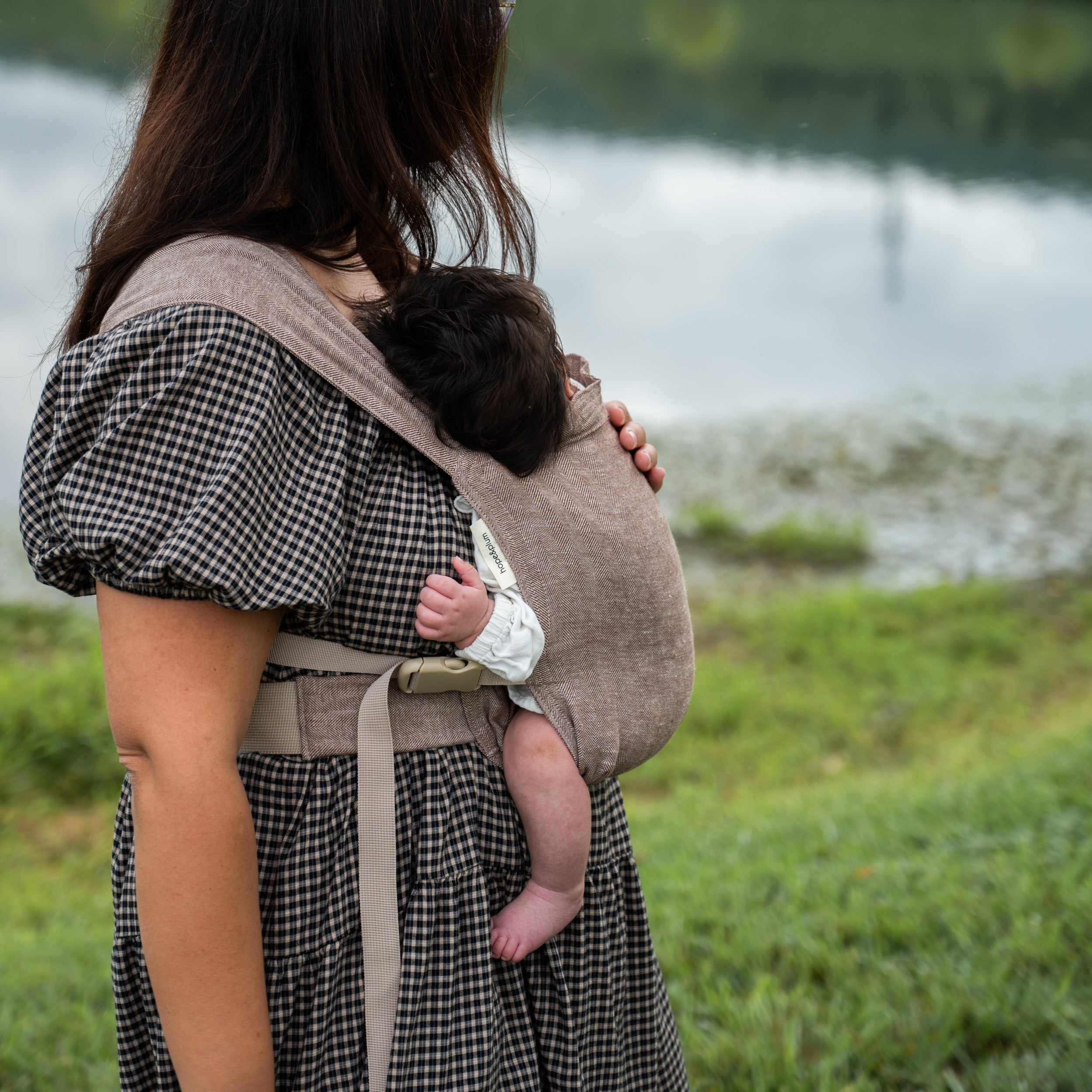 Woman in a gingham dress standing by a pond, carrying a sleeping newborn on her back in the Oak Sprout Newborn Carrier, a chocolate and oatmeal herringbone weave.