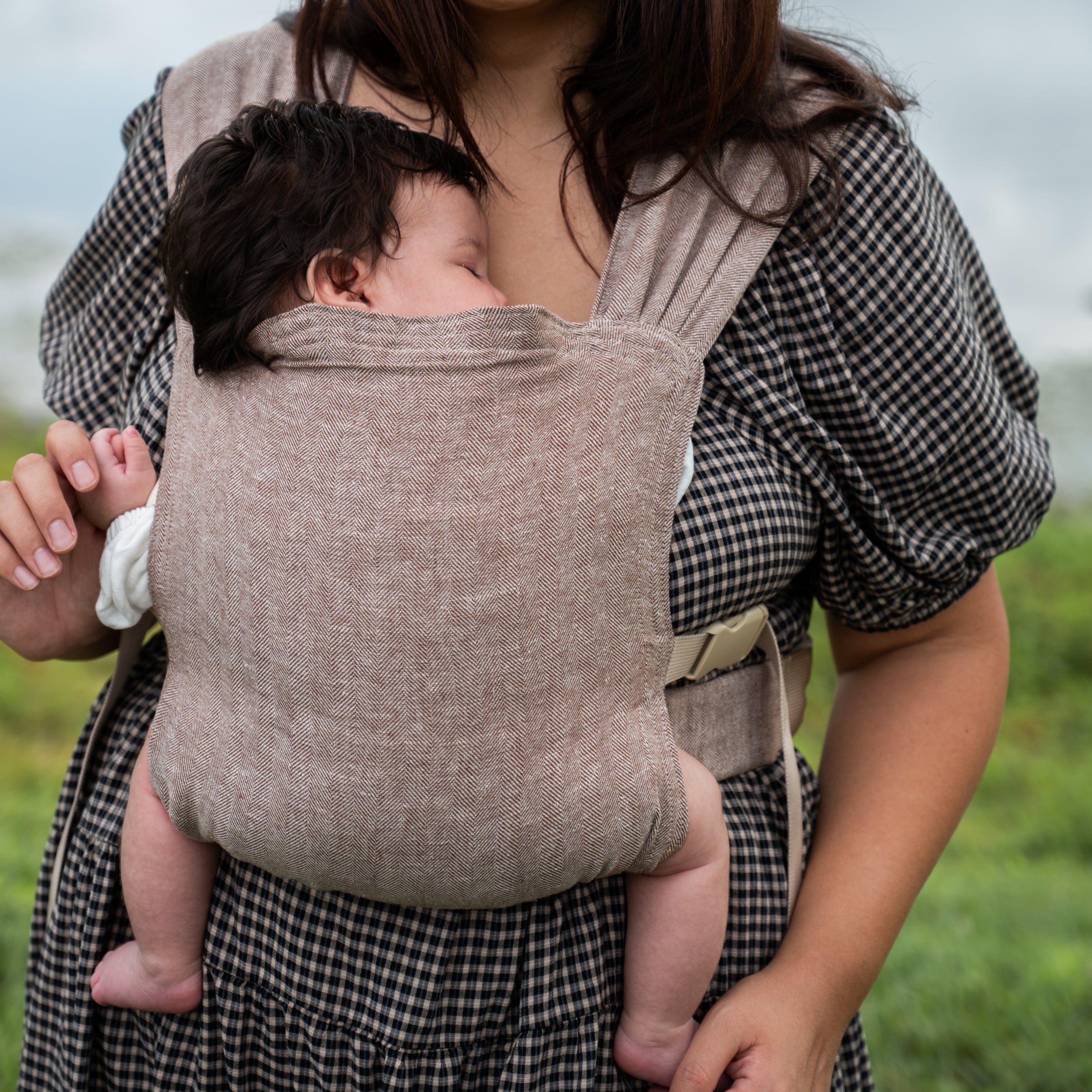 Close-up of a sleeping newborn worn on a woman's chest in the Oak Sprout Newborn Carrier, showing the chocolate and oatmeal herringbone weave and hope&plum label.