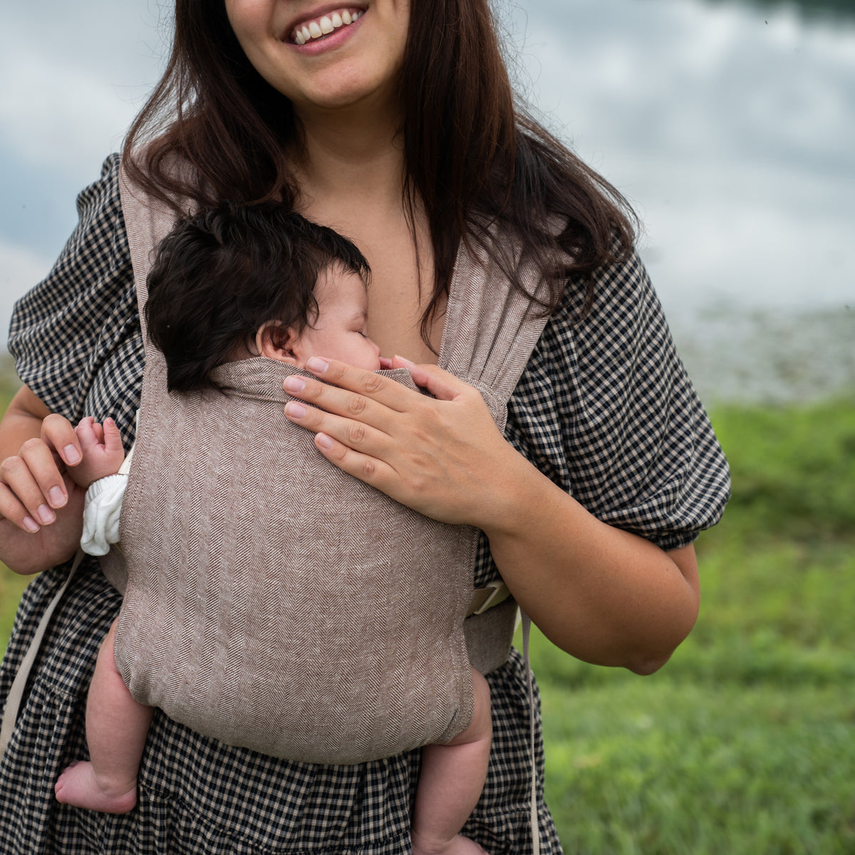Smiling woman holding a sleeping newborn against her chest in the Oak Sprout Newborn Carrier, a chocolate and oatmeal herringbone weave.