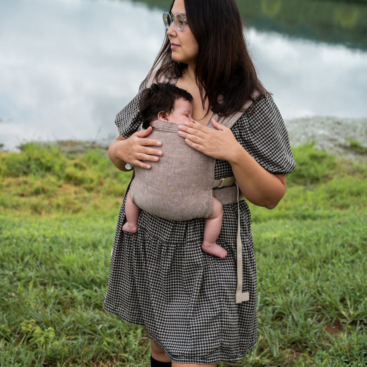 Woman with glasses looking down at a sleeping newborn worn on her chest in the Oak Sprout Newborn Carrier, a chocolate and oatmeal herringbone weave, standing by a lake.