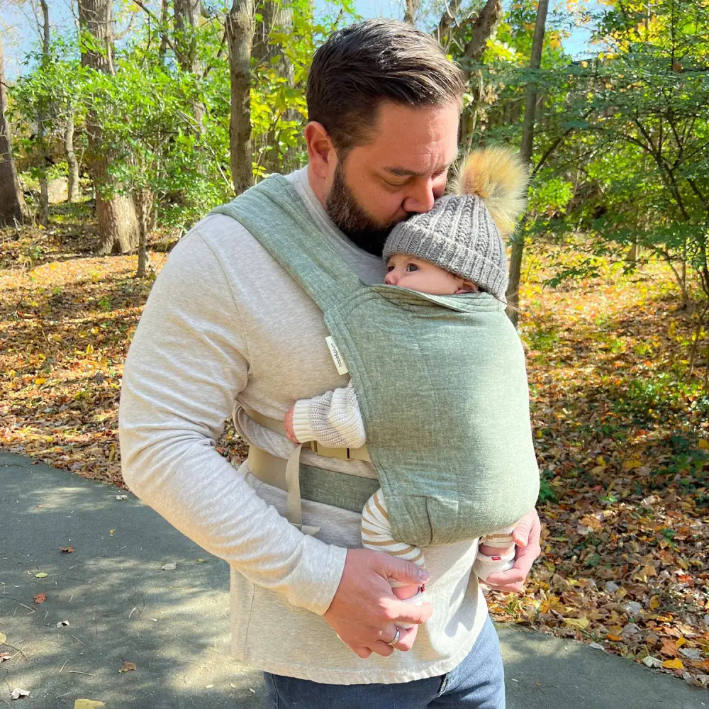 Father wearing infant in green baby carrier and kissing the baby's head.