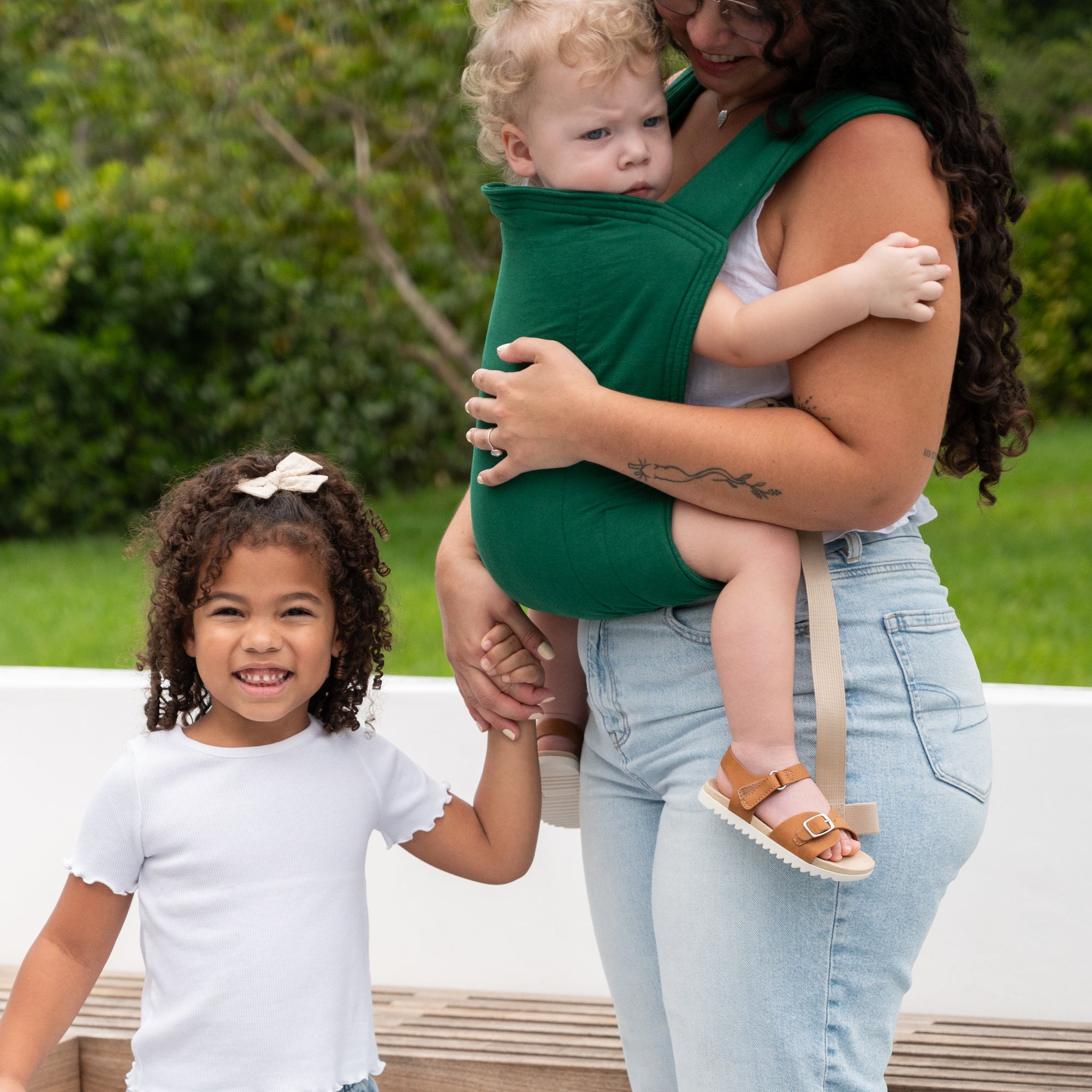 Woman holding a child in a green baby carrier with another child standing next to them outdoors.