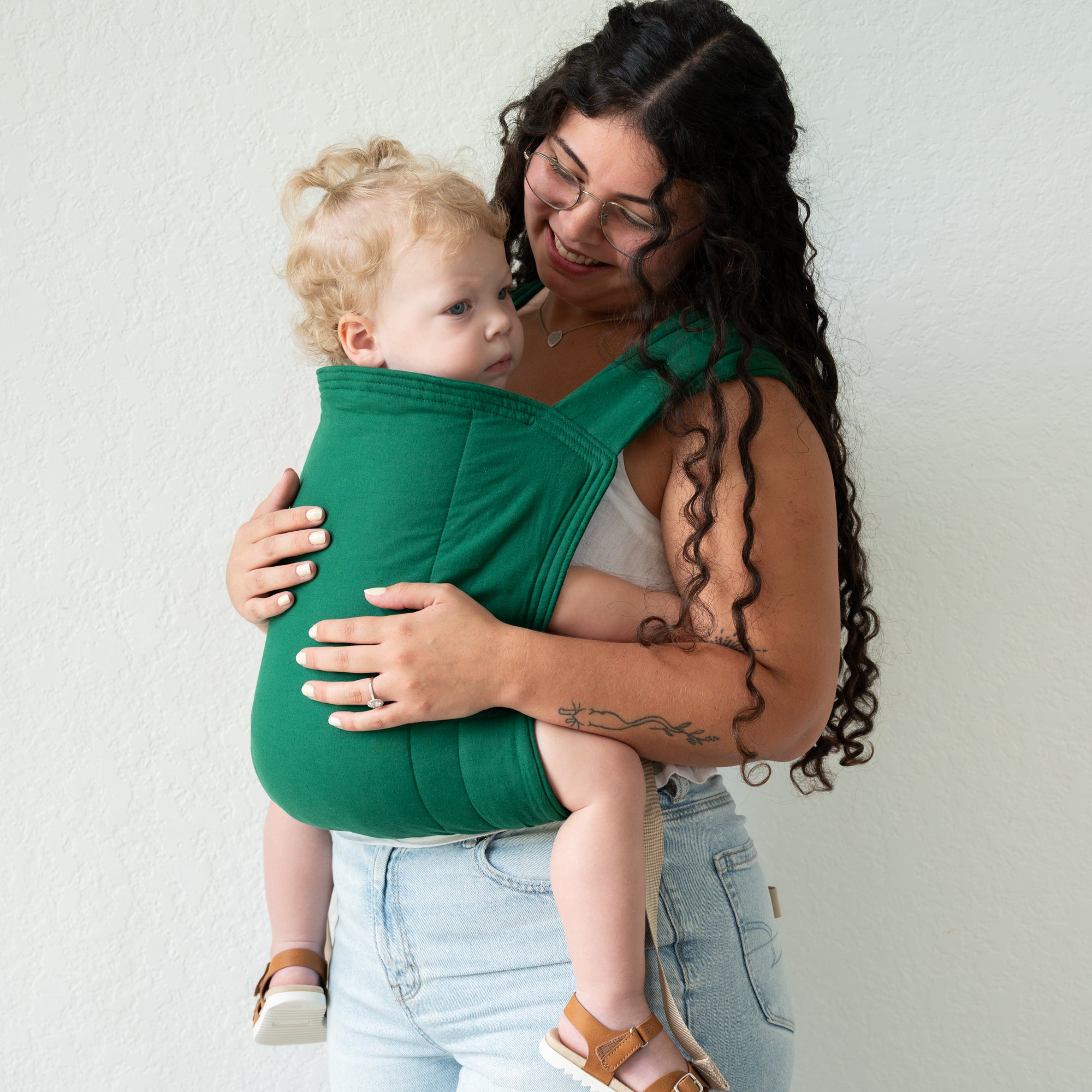 Woman holding a baby in a green carrier against a white wall
