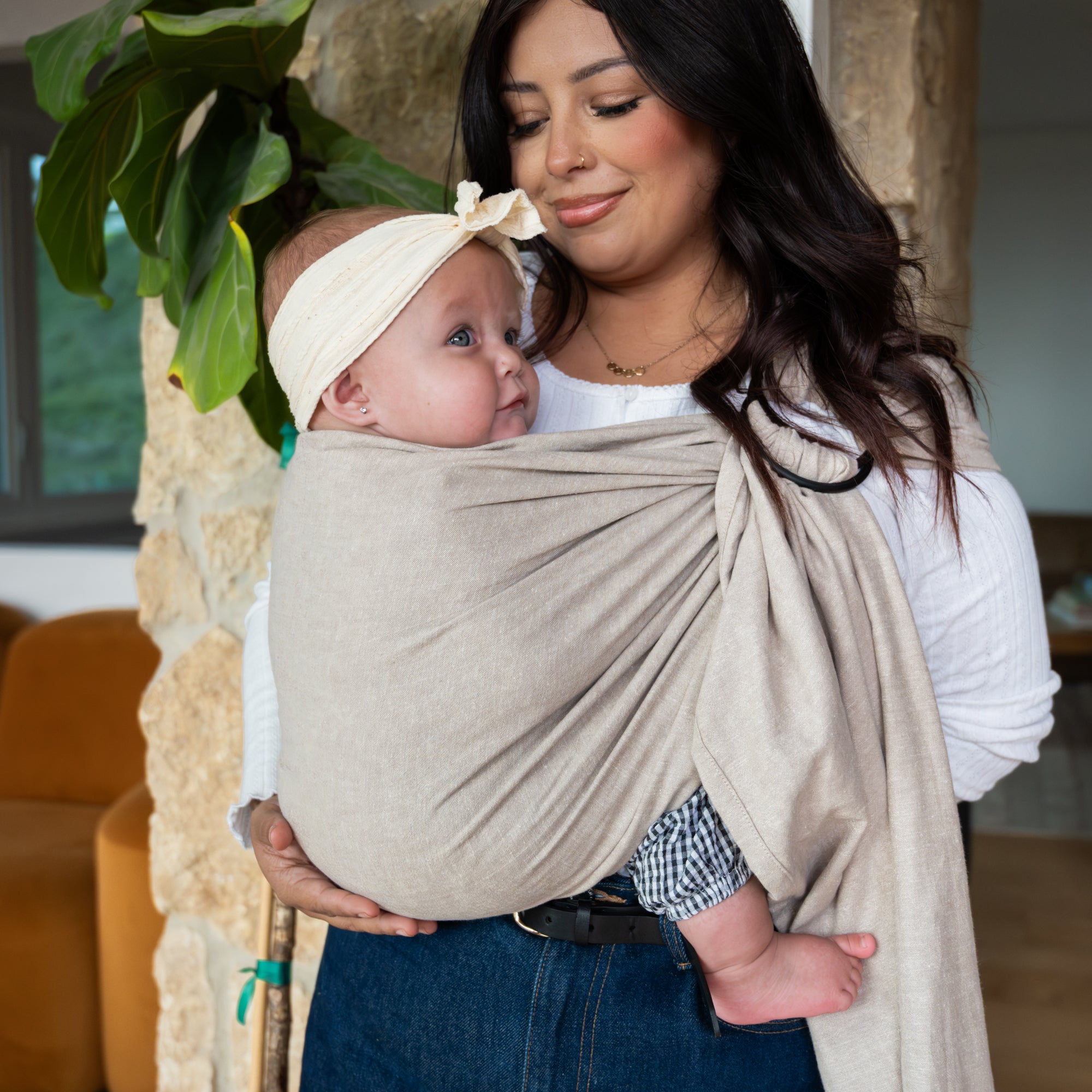 Woman holding a baby in a beige wrap carrier with a plant and stone wall in the background