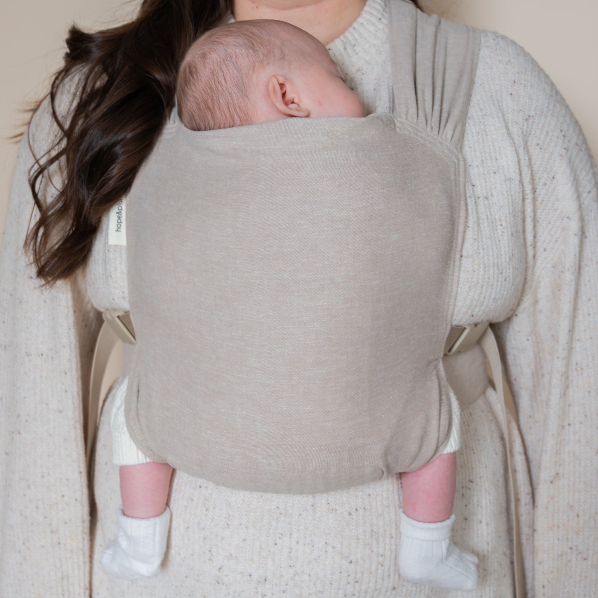 Person holding a baby in a beige baby carrier against a neutral background