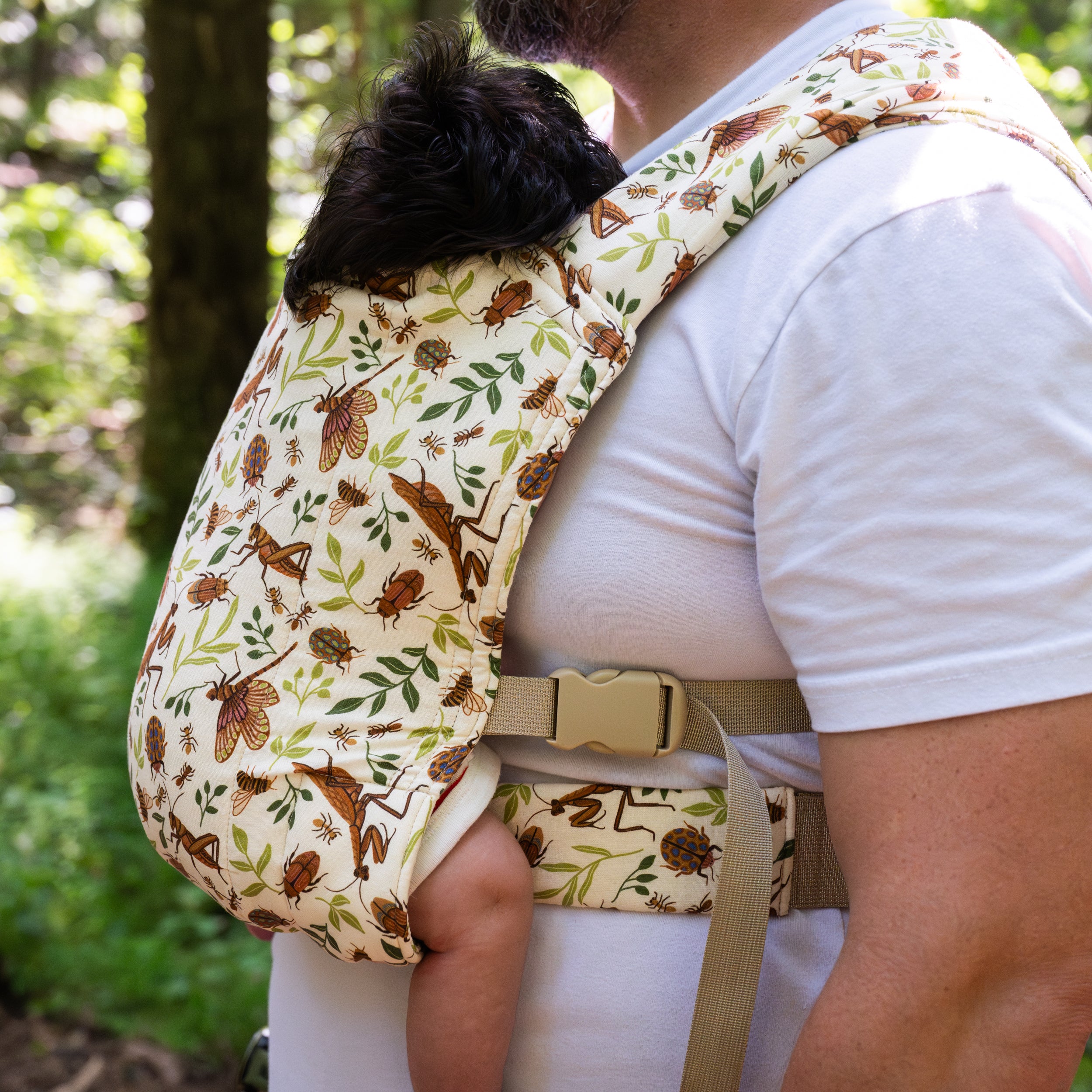 Close-up of a sleeping toddler worn on a man's chest in the Reed Lark Baby Carrier, a natural hemp-cotton fabric printed with warm brown bugs and leaves, showing ladybug, beetle, and dragonfly details.