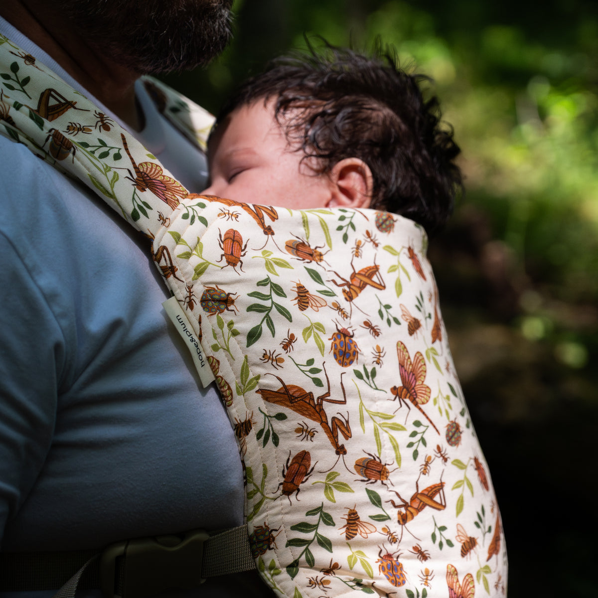 Close-up of a sleeping baby's face against a man's chest in the Reed Lark Baby Carrier, a natural hemp-cotton fabric printed with warm brown bugs and leaves.