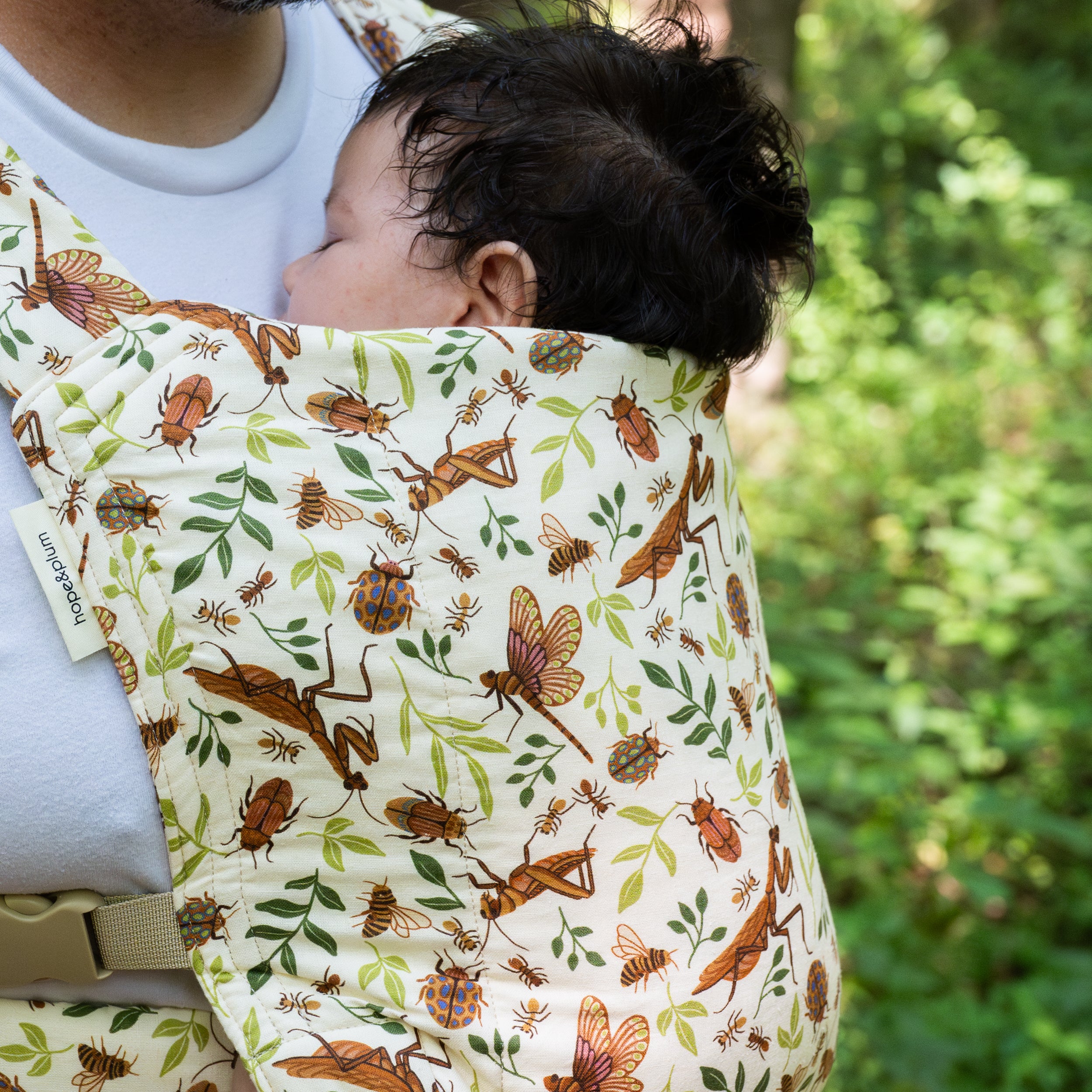 Close-up of a sleeping baby worn in the Reed Lark Baby Carrier, a natural hemp-cotton fabric printed with warm brown mantises, dragonflies, and beetles among leaves.