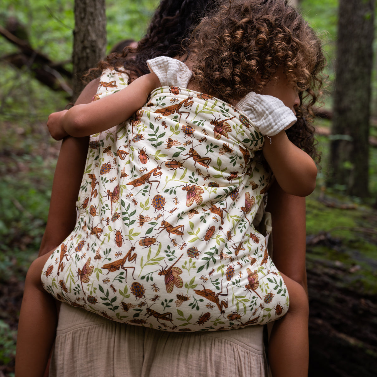 Back view of a mother carrying a toddler in the Reed Lark Kid Carrier, a natural hemp-cotton fabric printed with warm brown bugs and leaves, in a wooded setting.
