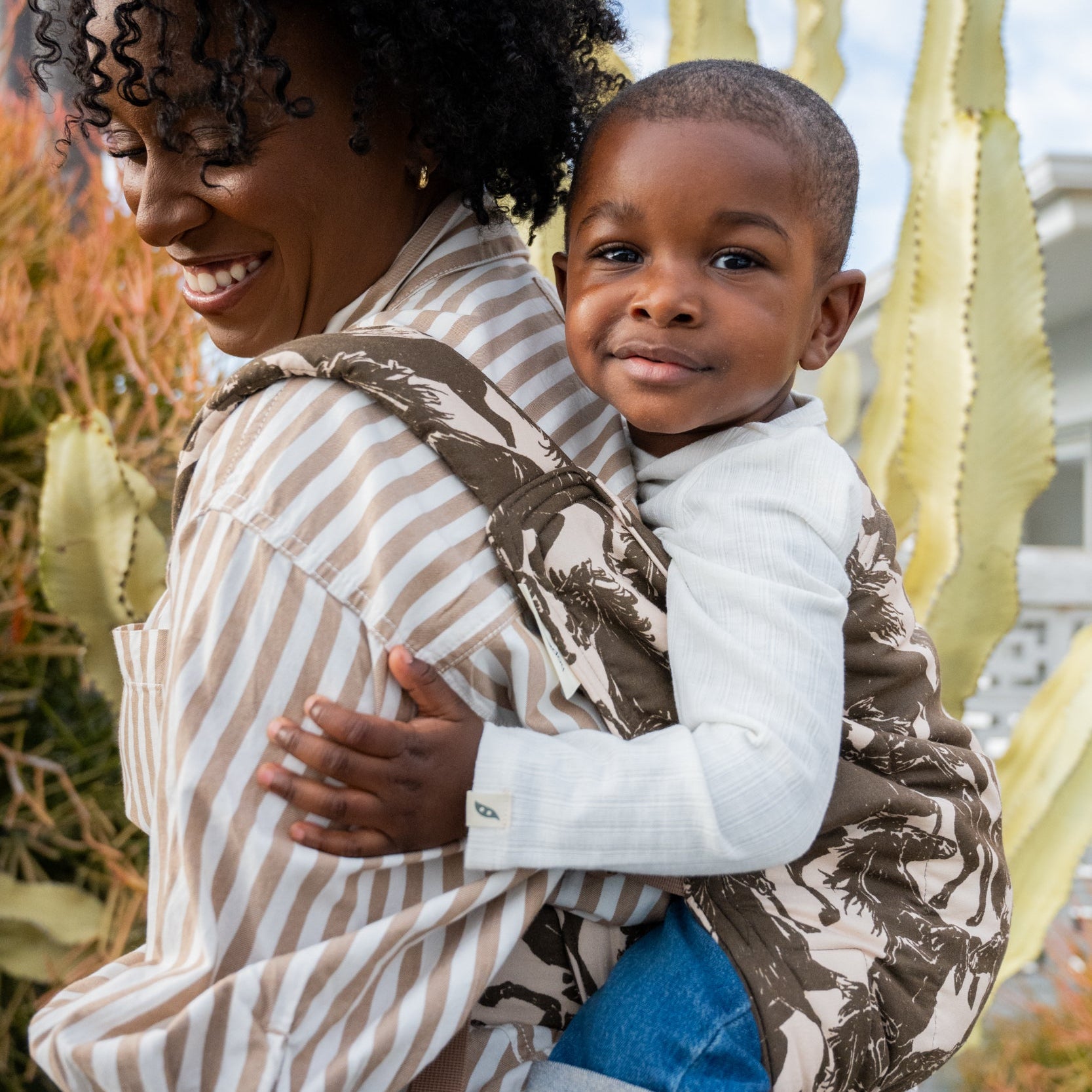 Lark Kid Carrier in Rodeo β caregiver carrying a smiling toddler on her back in the mustang print, cactus garden behind.