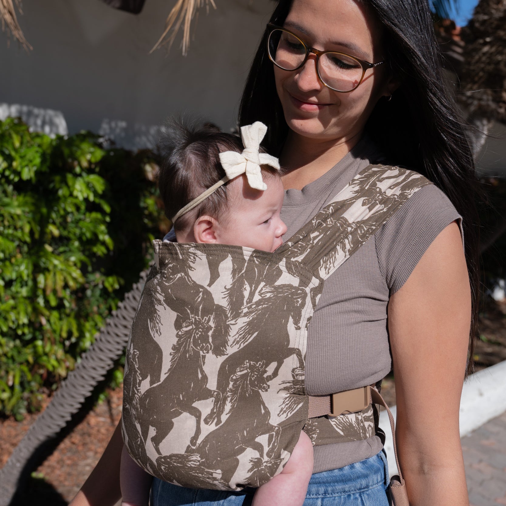 Sprout Newborn Carrier in Rodeo β caregiver walking with a newborn in the mustang print carrier, palm trees behind.