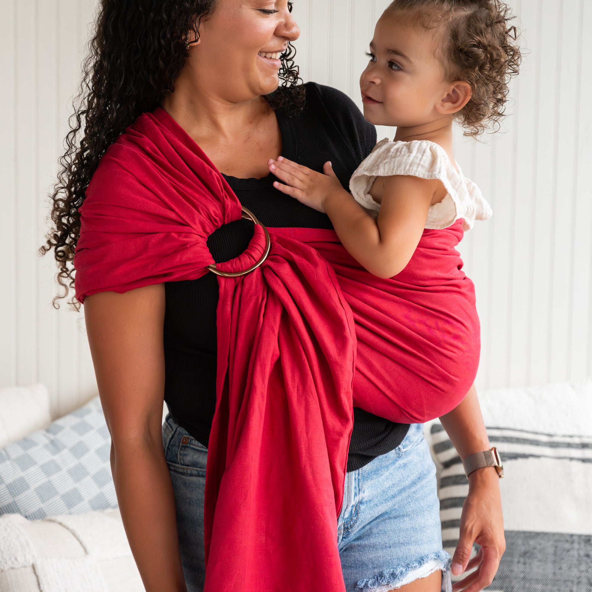 Woman holding a child in a red baby sling wrap, standing in a living room.
