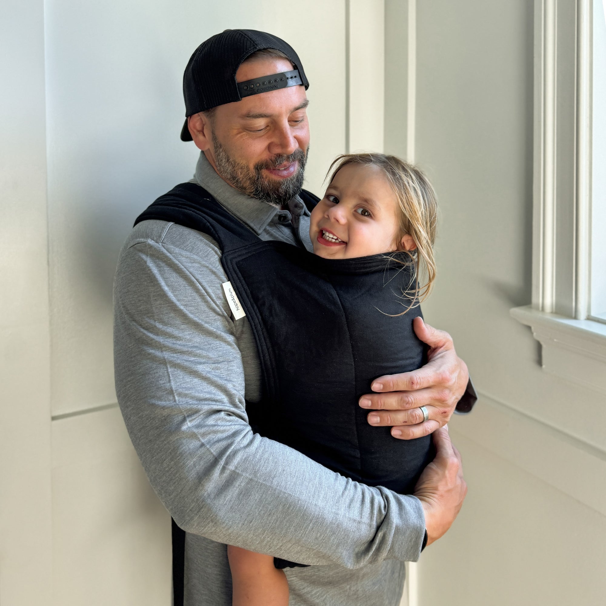 Man carrying a child in a black baby carrier against a light-colored wall.