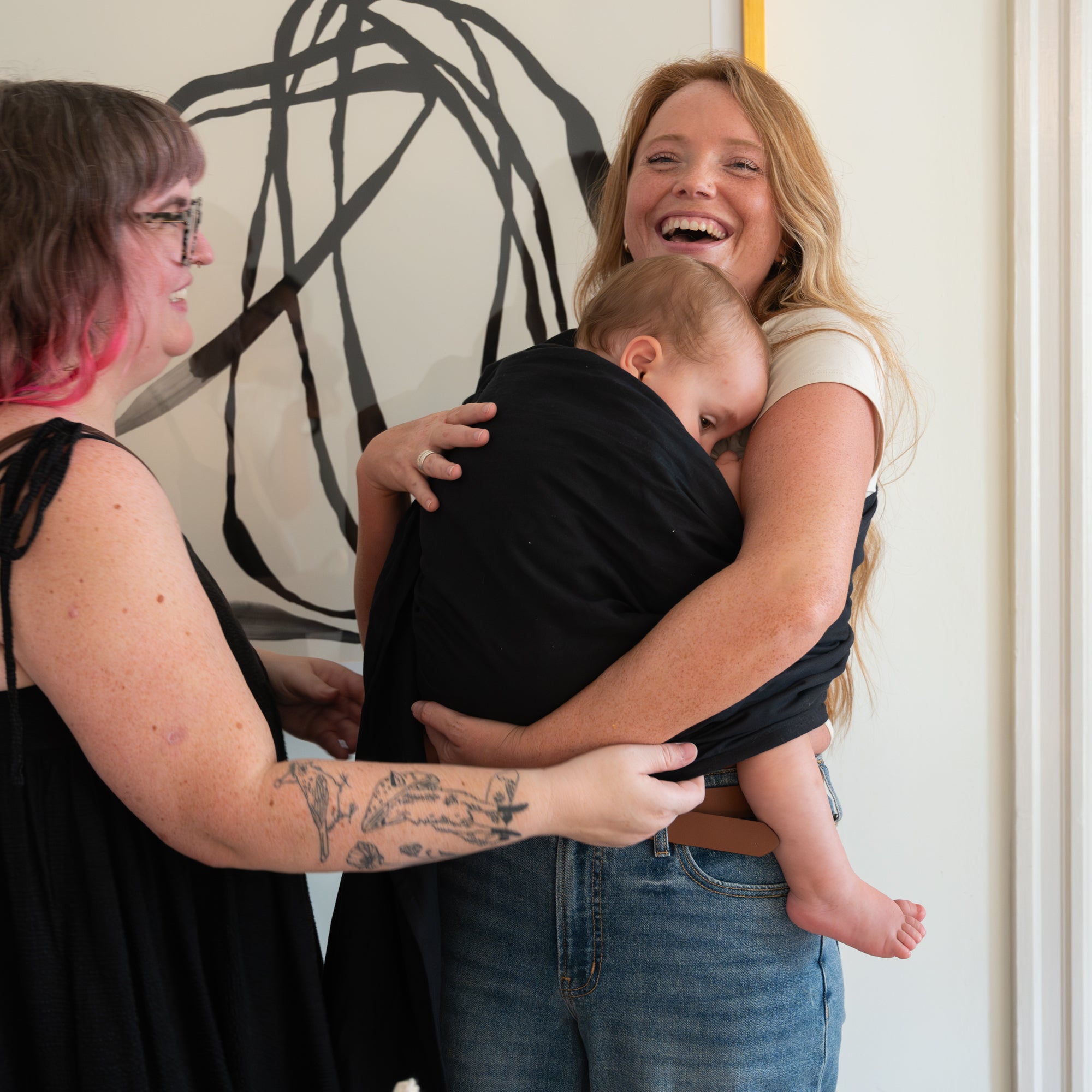 Two women holding a baby in a room with abstract art on the wall.