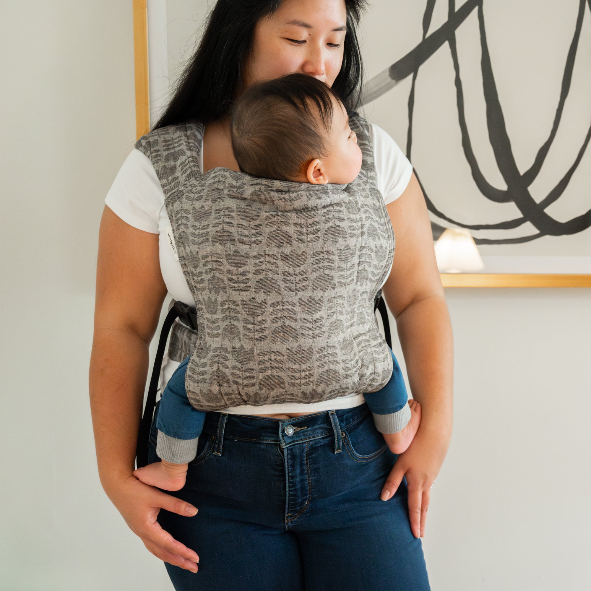 Woman holding a baby in a grey tulip patterned carrier against a neutral background