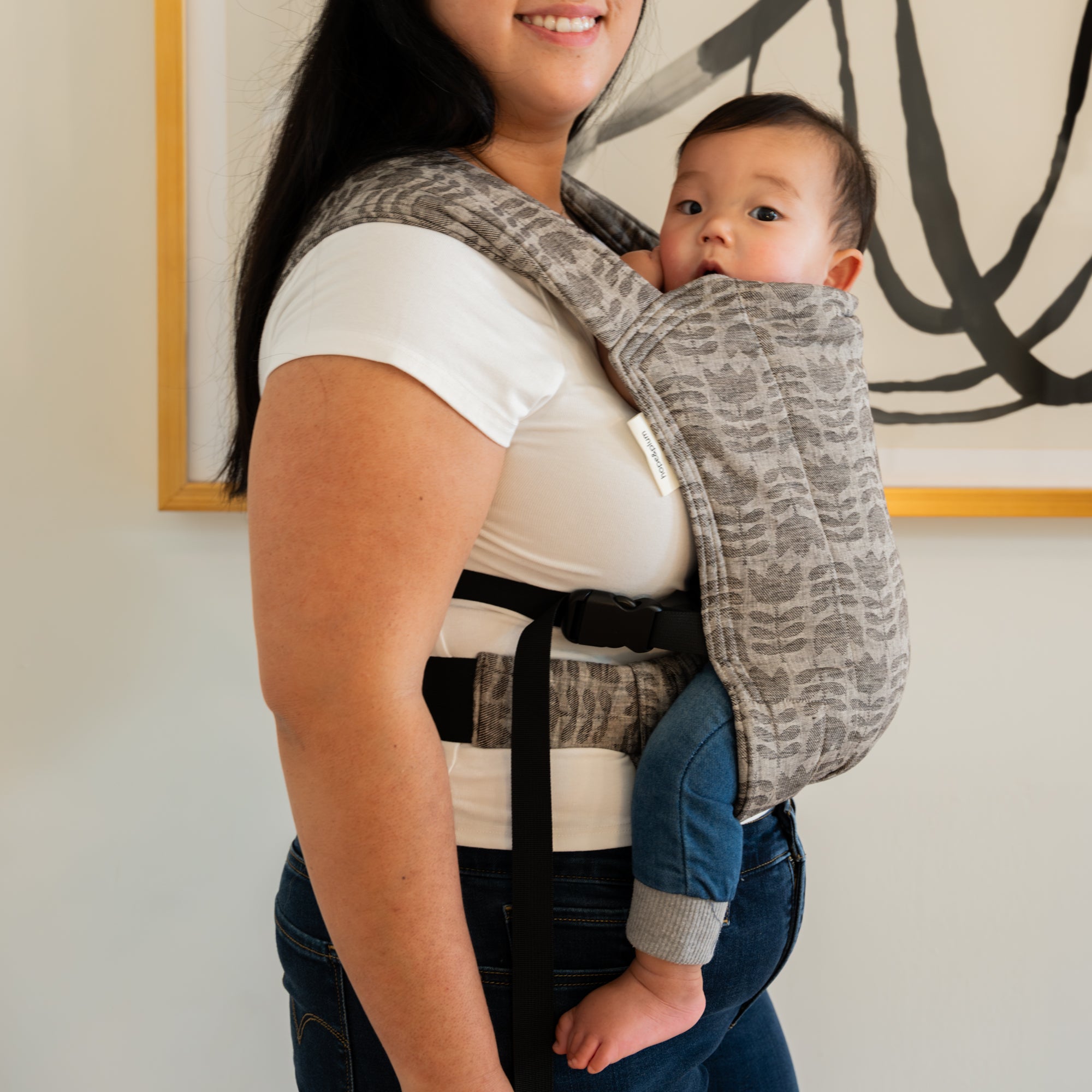 Woman carrying a baby in a gray tulip patterned baby carrier against a neutral wall.