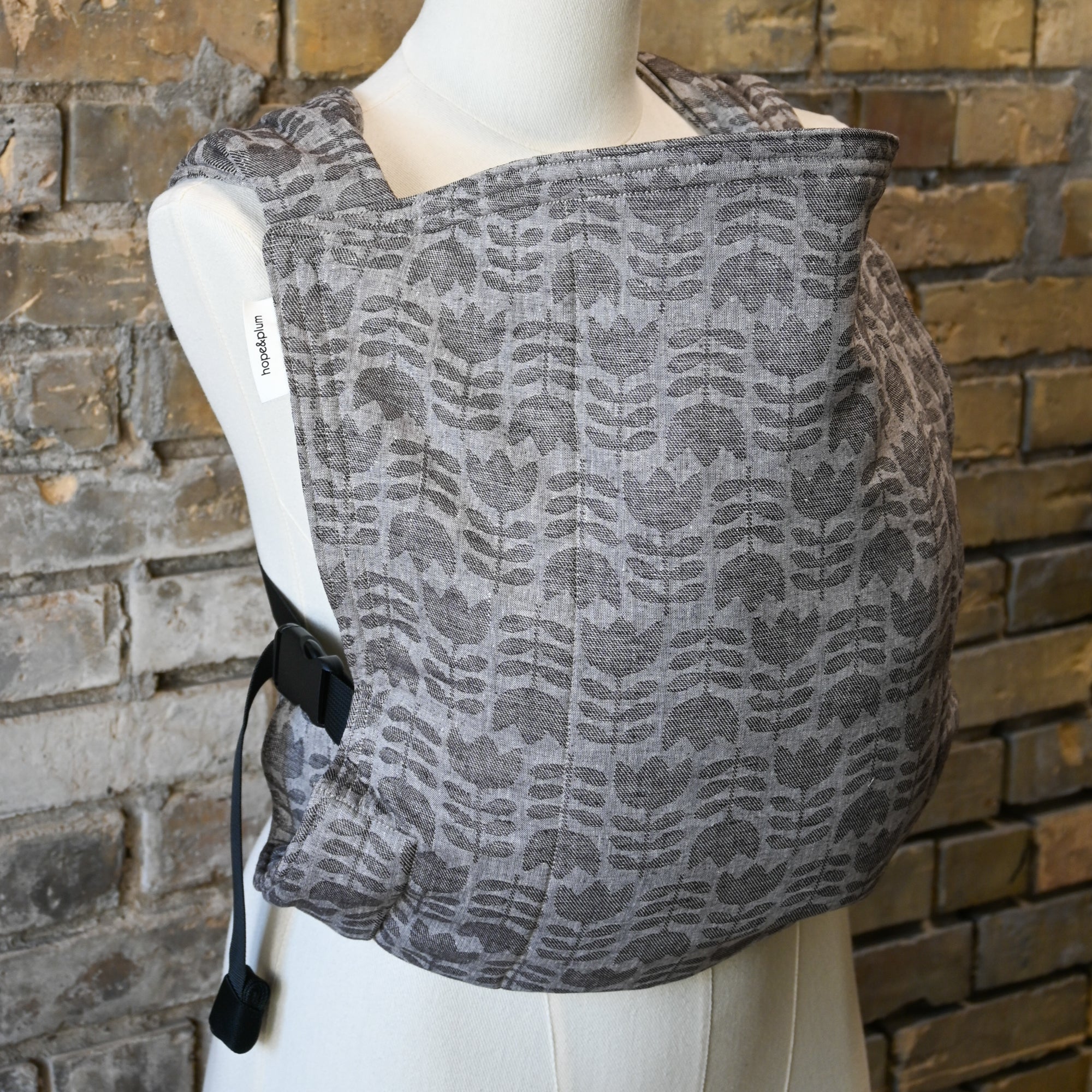 Gray tulip patterned baby carrier on a mannequin against a brick wall.
