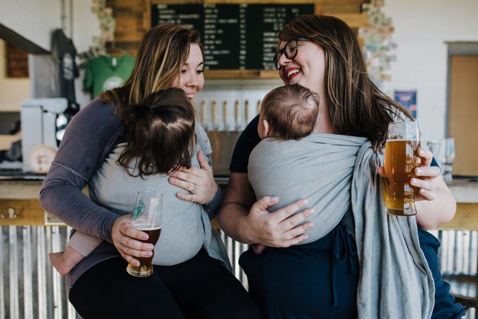Two woman, Skye and Mallory, wearing their babies in the same ring sling, Fin, a light blue color, while holding beers and smiling.